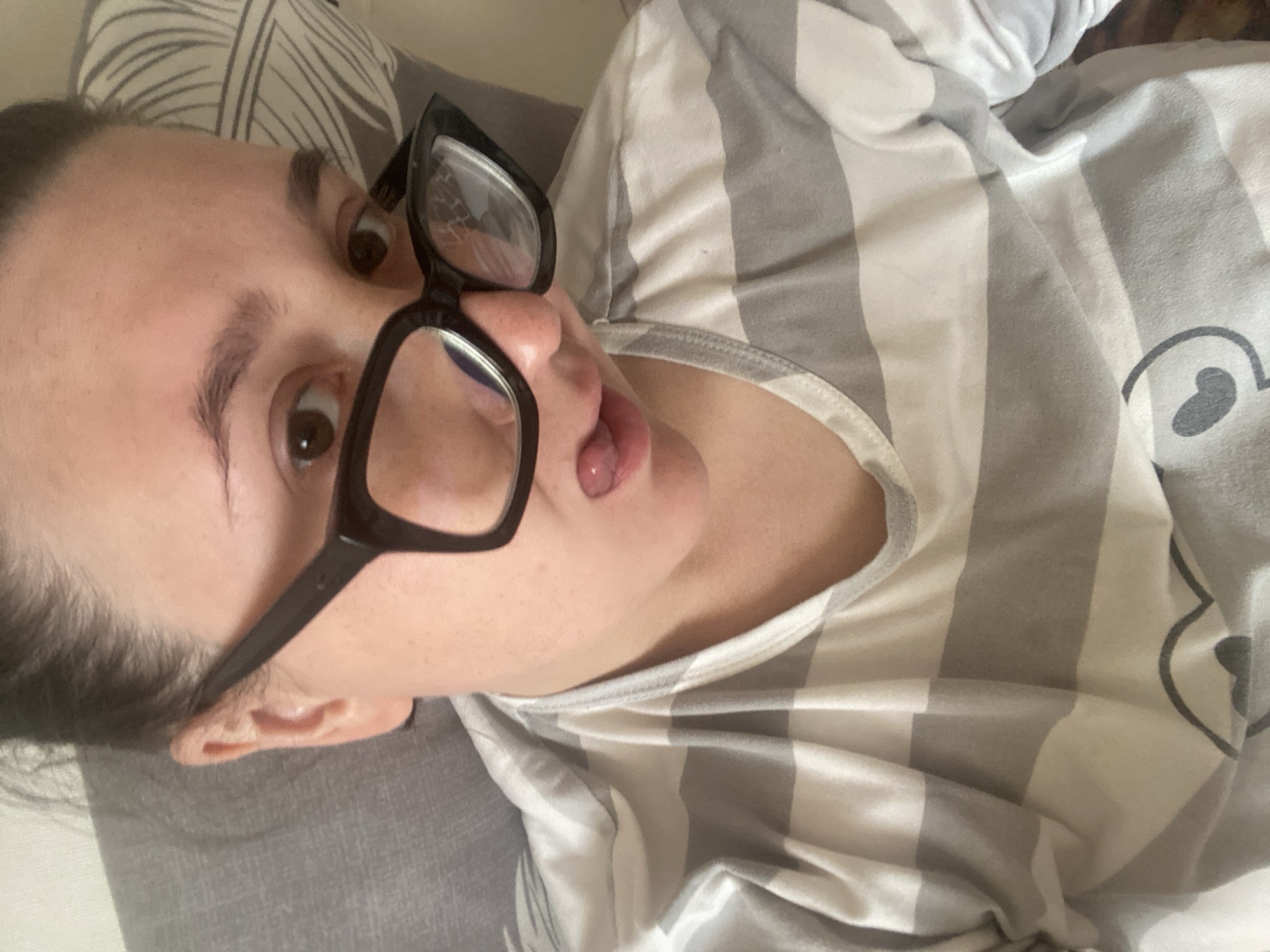 An indoor, close-up shot, likely taken in Chaikovsky, Russia, features a person lying down, looking towards the camera with a playful expression. The individual, whose gender is not explicitly identifiable, has wide, dark eyes and dark hair pulled back. They are wearing prominent black-framed eyeglasses that rest slightly on their nose. Their mouth is slightly open with their tongue gently sticking out, conveying a lighthearted and silly mood.
The person is dressed in a casual top or pajama shirt with alternating gray and white horizontal stripes. A partial graphic, resembling an outline of a bear or cartoon animal face, is visible on the striped fabric towards the right side of the frame. They appear to be resting on a bed or sofa, with a pillow featuring a subtle, light-colored leaf or feather pattern visible behind their head and to the left. The lighting is soft and ambient, consistent with an indoor setting during any time of day, emphasizing a relaxed and comfortable atmosphere. No text is visible in the image.