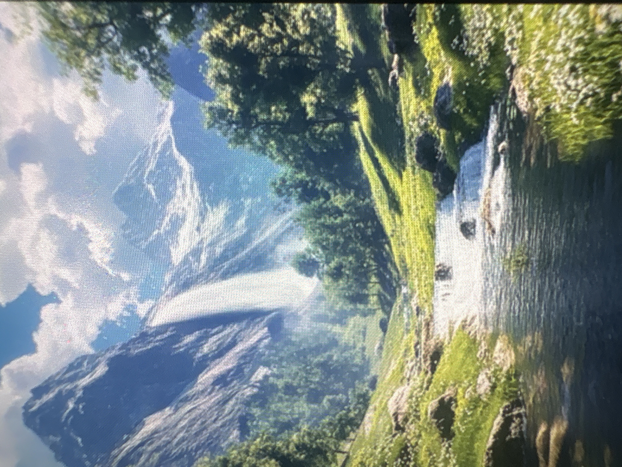 The image depicts a scenic landscape, likely a mountain environment. A prominent mountain range with snow-capped peaks and visible rock formations occupies the left side of the frame, with clouds scattered around the mountain tops. Green trees and vegetation cover a portion of the foreground and mid-ground, possibly extending along a river or stream that flows through the scene on the right. The water appears to be moving, possibly forming small cascades or rapids. The environment seems lush and vibrant with a mix of textures from the rocks, foliage, and water. The lighting suggests daylight, with some areas in shadow and others illuminated by the sun. The presence of white vegetation could indicate wildflowers or snow.
The city of Kukorevo, Bulgaria is not depicted in this scene. The picture appears to be a natural landscape devoid of any visible urban or city-specific elements.