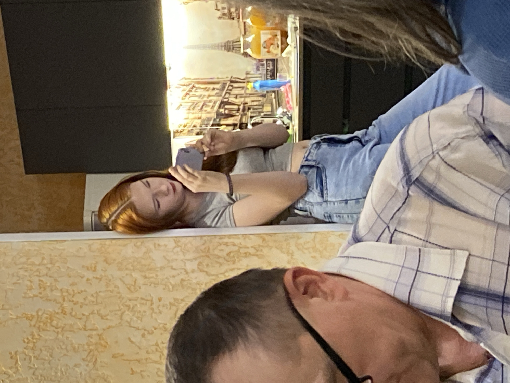 A woman with auburn hair, wearing a gray t-shirt and blue denim shorts, is lying on a surface, holding a smartphone. An image depicting the Eiffel Tower is visible in the background. A person with short gray hair and glasses, wearing a plaid shirt, is positioned in the foreground. The location is possibly Chaikovsky, Russia.