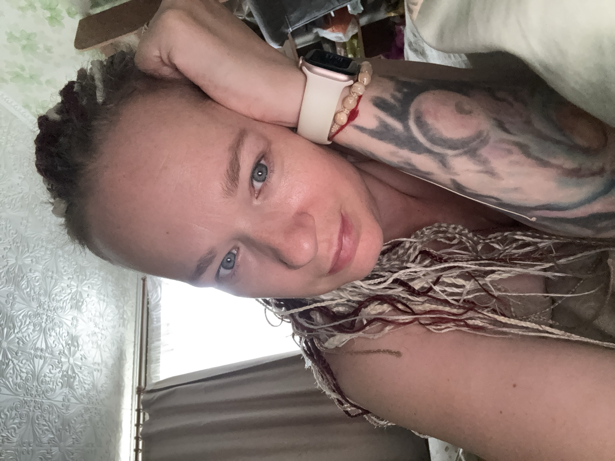 The media file contains a close-up, sideways portrait of a person with light skin, blue eyes, and braided hair featuring streaks of red and blonde. The individual is resting their head on their hand, with their arm also visible. They are wearing a light-colored smartwatch with a pink-gold casing and a white band on their left wrist. A beaded bracelet with red string accents is also worn on the same wrist. A large tattoo is visible on the individual's upper arm, featuring swirling patterns and shades of grey and black.
The background is a textured wallpaper with a floral pattern in a light, possibly greenish-white color. To the right of the person, a glimpse of a window with blinds and what appears to be a piece of furniture is visible, suggesting an indoor setting. The overall lighting appears to be natural, possibly indicating daytime. The individual's expression is relaxed, with a slight smile. The image appears to be a selfie, taken from a low angle. There is no visible text in the media file. The location context is Moscow, Russia.