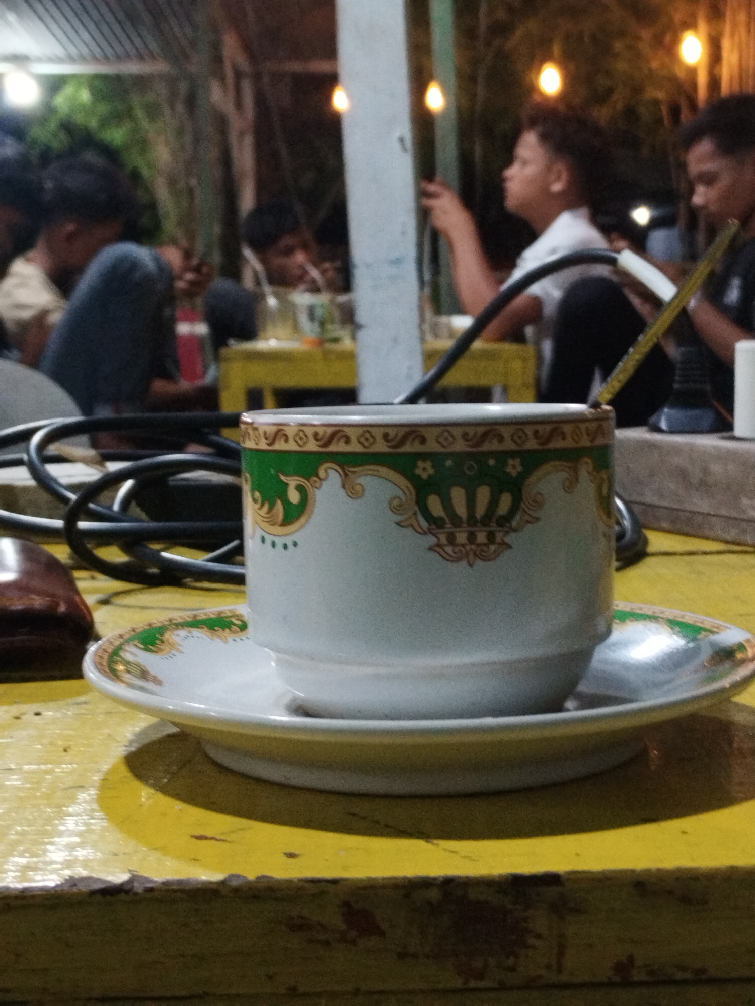 The image captures a scene likely within Lhokseumawe, Indonesia, based on cultural context. In the foreground, a white ceramic teacup sits on a saucer, both adorned with a green and gold crown-like pattern. A yellow spoon is resting in the teacup. The table underneath is painted yellow and appears worn.
In the background, a group of people, primarily young males, are visible. They appear to be casually socializing, possibly enjoying beverages or engaging with electronic devices. Some are seated at yellow tables. Overhead lighting illuminates the scene. Electrical cables are present on the table in the foreground, near the teacup. The environment appears to be a cafe or outdoor seating area.