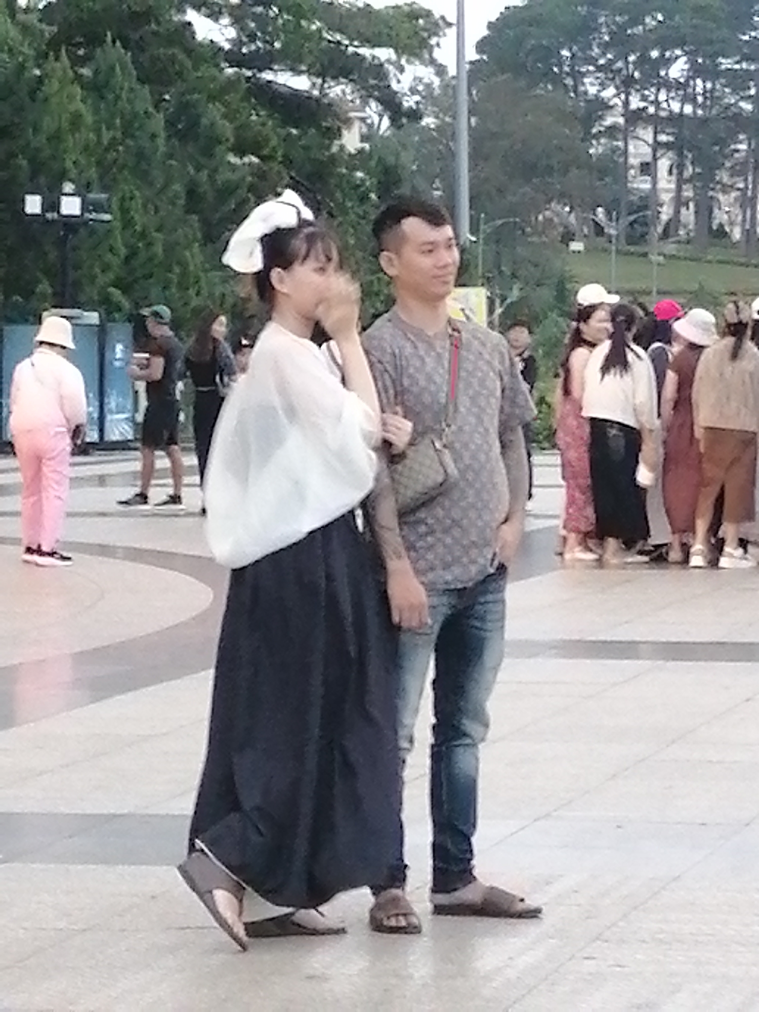 In Da Lat, Vietnam, two individuals stand prominently in the foreground of a public outdoor space. The male subject on the right, wearing a grey patterned t-shirt and blue jeans, has his left hand in his pocket and is looking to his left. He carries a small, cross-body bag. To his left, a female subject, wearing a white translucent overlay over a dark skirt and a large white bow in her hair, is turned slightly away, her face obscured as she brings her hands to her mouth. Her attire suggests a cultural influence. Behind them, a diverse group of people are visible, some wearing hats and casual clothing, suggesting a tourist or leisurely gathering. In the background, a line of trees with dense foliage borders the area, with some distant buildings peeking through. The ground is paved with light-colored tiles, some of which are arranged in a circular pattern, indicative of a plaza or walkway. The lighting suggests it is daytime, with the sun casting soft shadows.