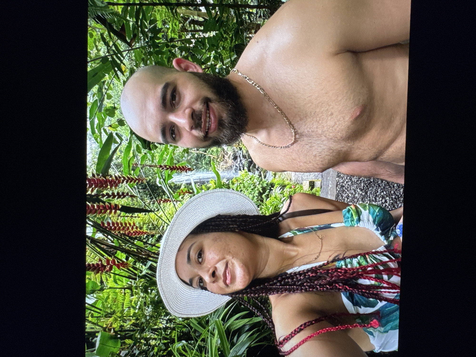 A selfie shows a man and a woman posing for a photo outdoors in the city of Unknown, Unknown. The man is shirtless and has a bald head, dark beard, and gold chain. The woman is wearing a white hat and a floral print top, and has long braids with red accents. They are standing in a lush, green environment with various plants and flowers. A small waterfall can be seen in the background.