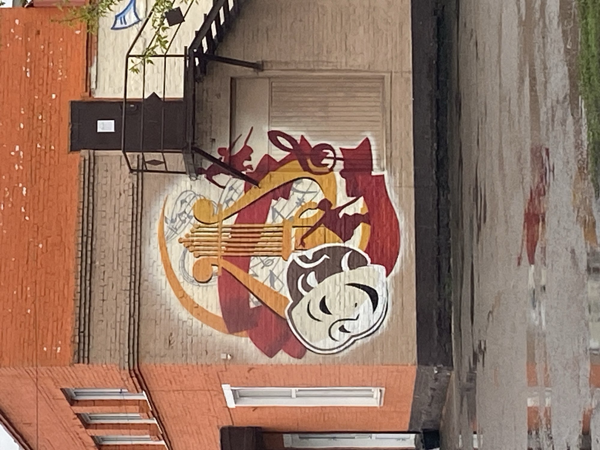 The image shows a brick building exterior with a large mural. The mural is a circular design containing musical and theatrical symbols. These include a lyre, a treble clef, and theatrical masks, painted in shades of gold, red, and white. The building has architectural details such as windows and a fire escape. The mural is painted on a beige brick wall, and the building has different sections of brick in orange. The environment suggests an urban setting. The location is Novomaltinsk, Russia.
