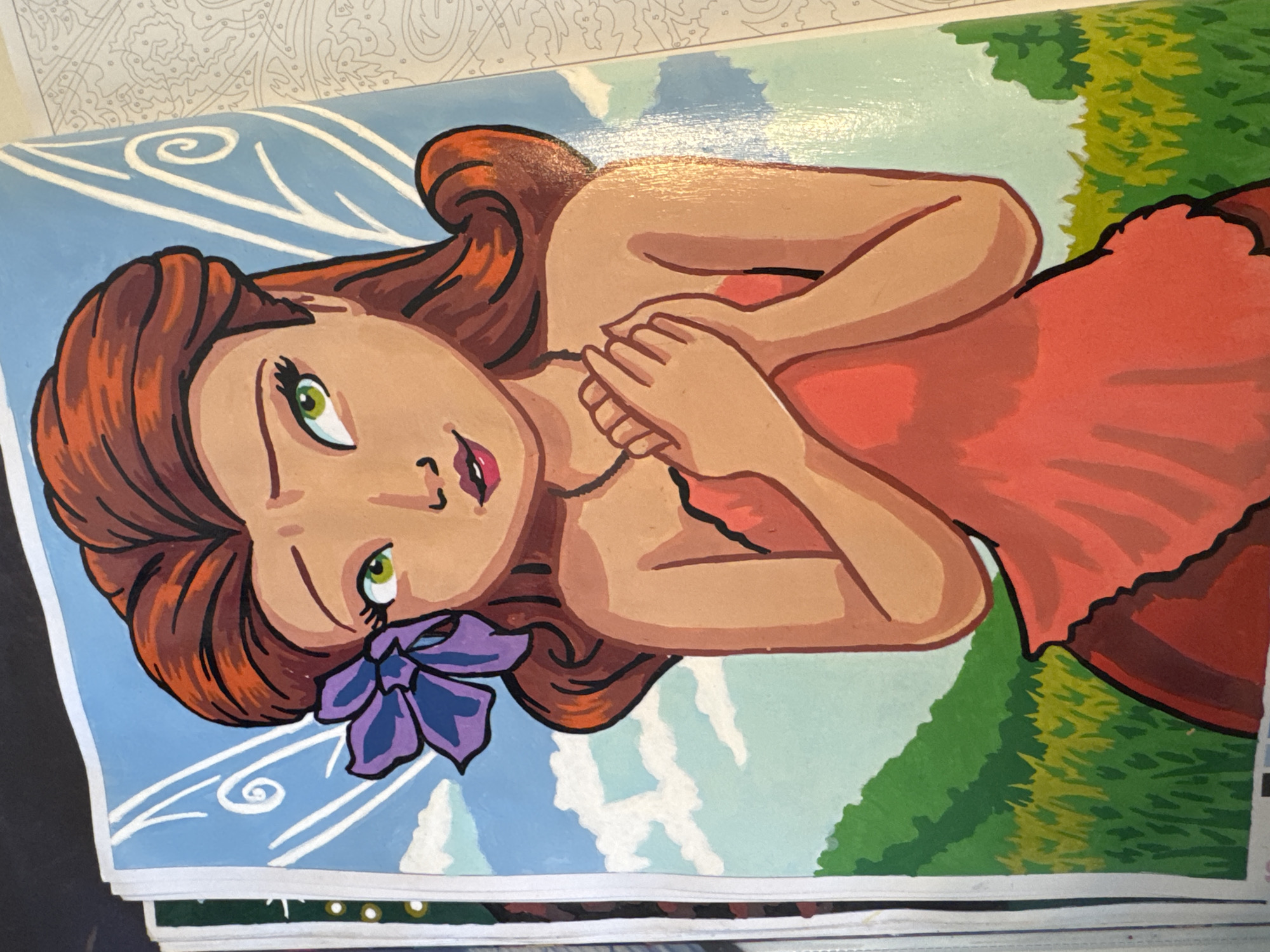 A close-up, eye-level, full shot captures a painted illustration of a fairy with auburn hair, green eyes, and a purple flower tucked behind her ear, against a backdrop of a blue sky with white clouds, and a green landscape. The fairy is depicted from the chest up, wearing an orange dress, with her hands clasped over her chest. Her head is tilted slightly to her right, and she has a coy expression. The illustration appears to be part of a paint-by-numbers book, as faint numbers are visible on the right edge of the image, suggesting an unfinished project. The lighting is bright, and the colors are vibrant. The fairy's wings are visible behind her head, rendered in white with subtle swirling designs.
