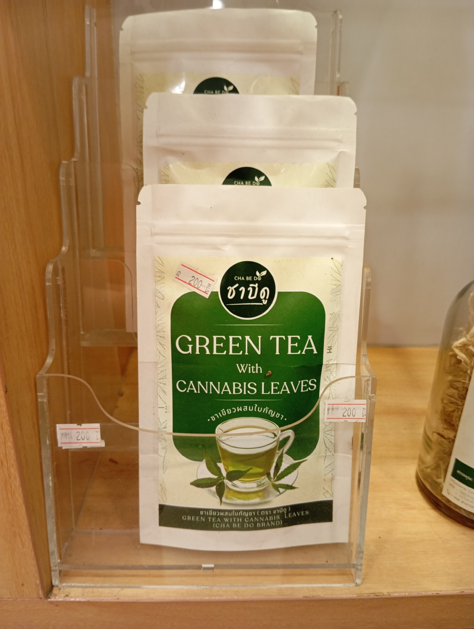 In Karon, Thailand, a clear acrylic display stand holds multiple pouches of green tea with cannabis leaves. The pouches are stacked vertically, with the front pouch prominently displaying the brand "CHA BE DO" and text in both Thai and English. The English text reads "GREEN TEA With CANNABIS LEAVES". A price tag of "200" is attached to the front of the display stand and also on the front of the pouch. The background consists of a wooden surface and a blurred glass jar containing dried plant material. The scene is well-lit, suggesting an indoor retail environment, possibly a shop or market stall.