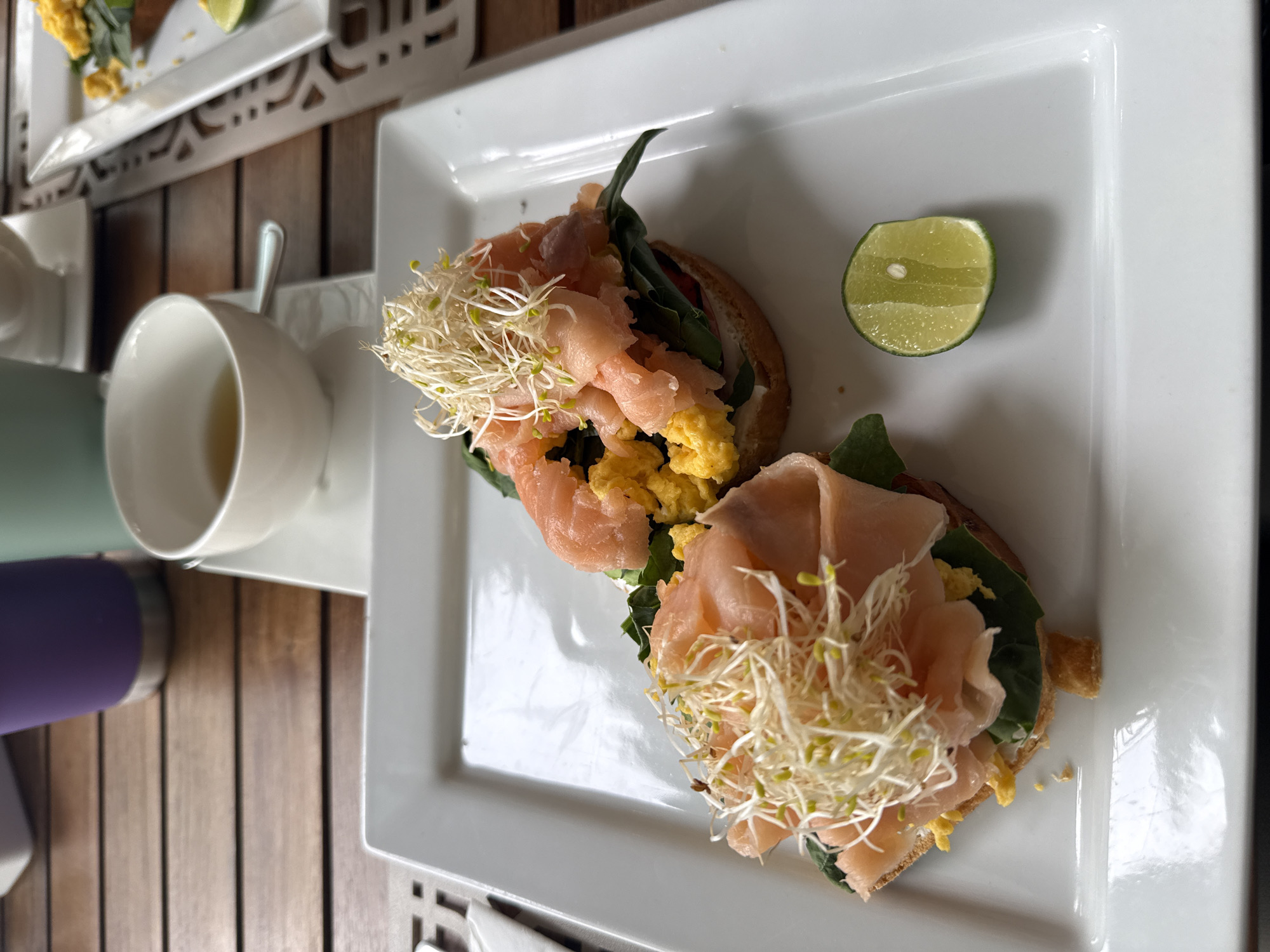 On a wooden surface in Huixquilucan de Degollado, Mexico, there is a rectangular white plate holding two open-faced sandwiches. Each sandwich is topped with sprouts, light pink sliced fish, spinach, and scrambled eggs, and is accompanied by a half lime. Next to the plate is a white cup with a silver spoon on a white saucer, along with the base of a purple tumbler. Another plate with a similar sandwich is partially visible in the background.