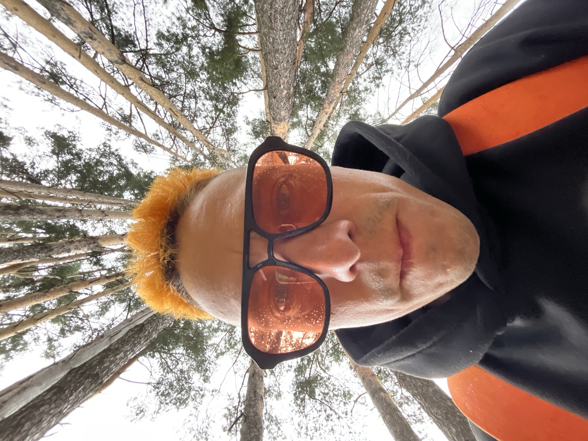 An upside-down, close-up, low-angle selfie captures a person wearing orange-tinted sunglasses with a black frame. The person has bright orange hair and is wearing a black hooded garment with an orange stripe visible across the chest. The background consists of tall trees, appearing to be pine trees, with a bright, overcast sky. The scene suggests an outdoor setting, possibly a forest or park. The image is sharp and well-lit, with the focus primarily on the person's face and upper body. The reflection in the sunglasses shows more trees and the sky. The overall impression is that of a candid, spontaneous shot taken in a natural environment.