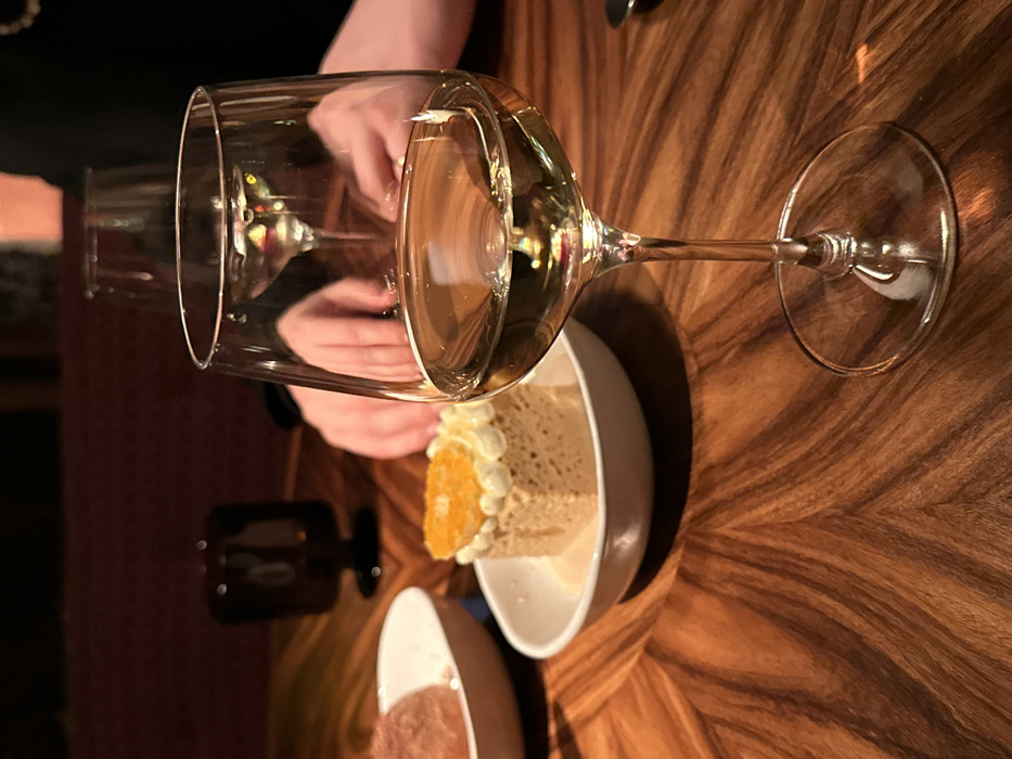 A tilted, overhead shot features a wine glass filled with white wine, partially overlapping a dessert plate containing a slice of cake. The cake is adorned with white frosting and a slice of orange. The setting is on a wooden table, showcasing the table's unique grain pattern. Another small dish containing a pink dessert is partially visible at the bottom of the image. A second wine glass appears blurred in the background.