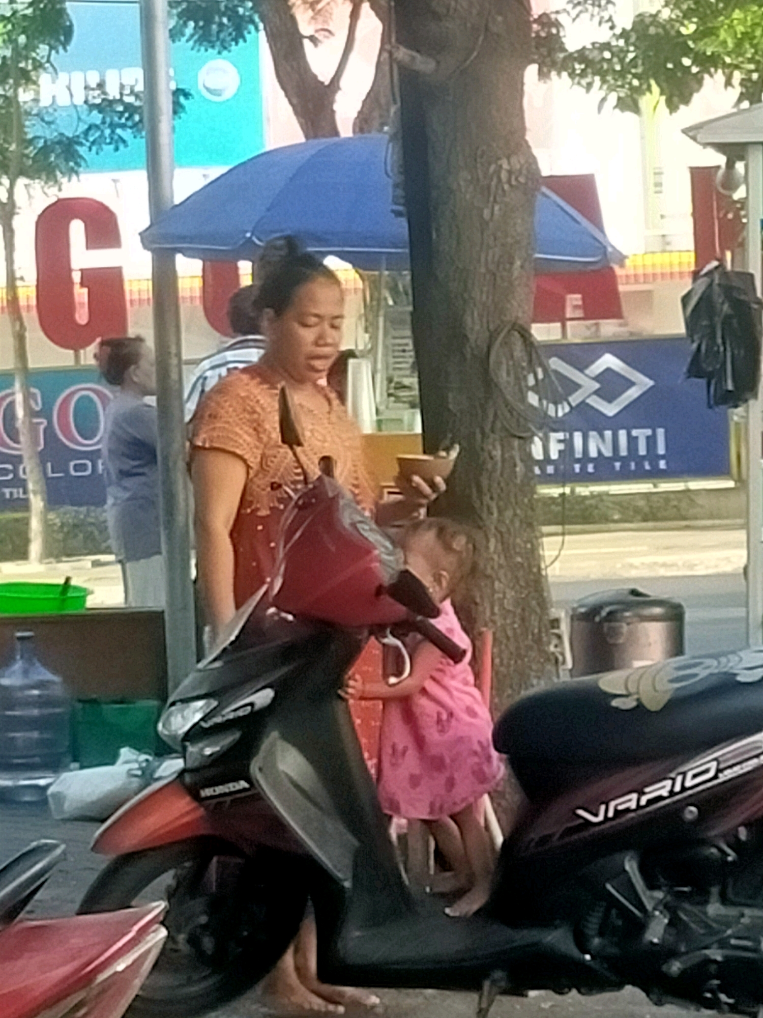The image depicts a street scene in Surabaya, Indonesia, near a commercial area. In the foreground, a woman in an orange patterned blouse is holding a small child in a pink dress. The woman appears to be speaking or calling out. The child is embracing the woman. They are positioned next to a red and black motorcycle. Behind them, a large tree trunk is visible. In the background, a blue umbrella is open, and signs with lettering, including "GO COLOR" and "FINITI TILE," are displayed on buildings. Water bottles and green containers are also present. The lighting suggests it is daytime.