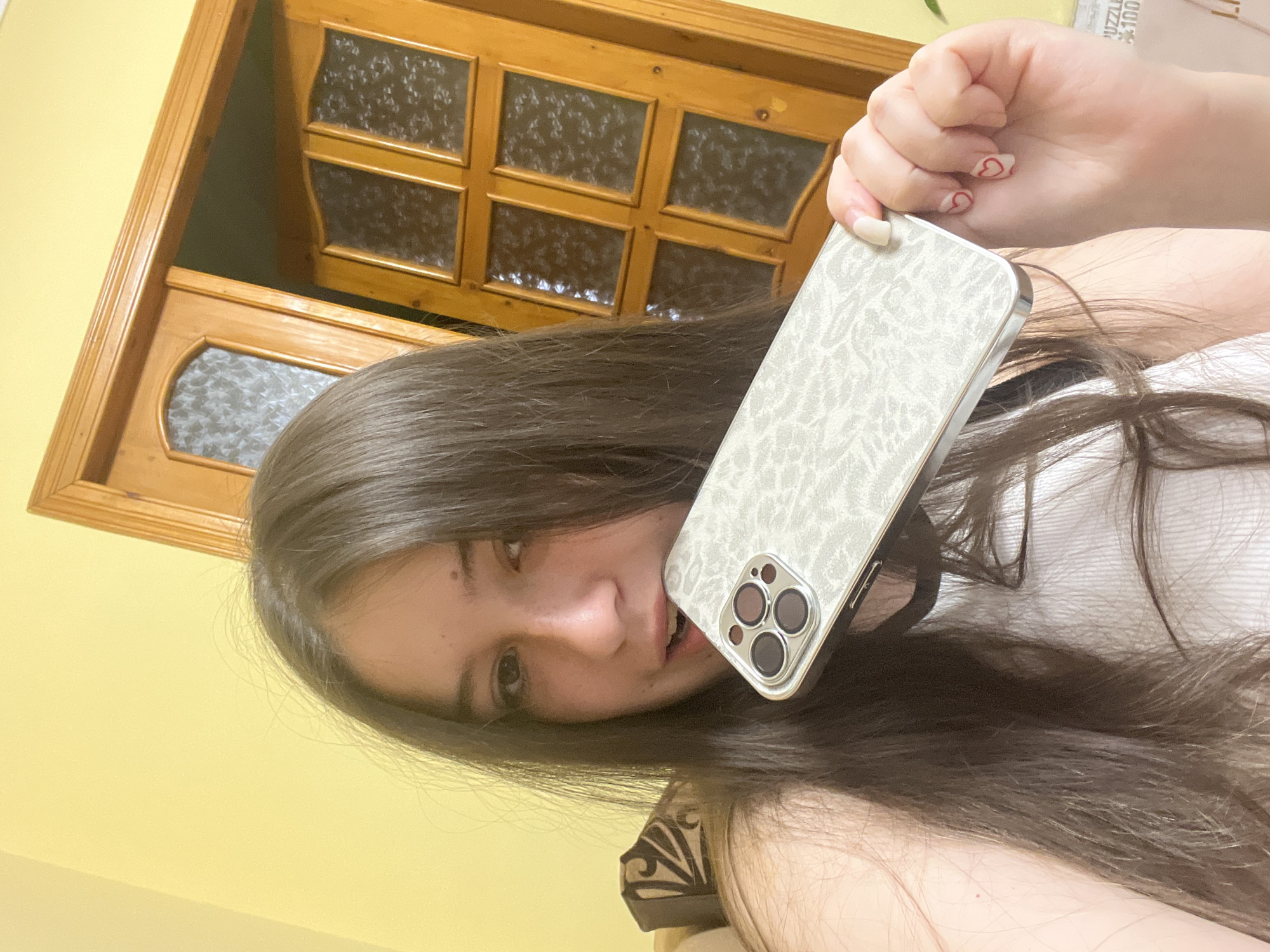 A woman with long brown hair is holding a smartphone up to her face, partially obscuring her mouth. The phone has a white patterned case with three visible camera lenses. She has painted fingernails with small red hearts on them. The background includes a yellow wall and a wooden-framed door with decorative glass panels. There is no information available indicating the location is Cherliany, Ukraine.