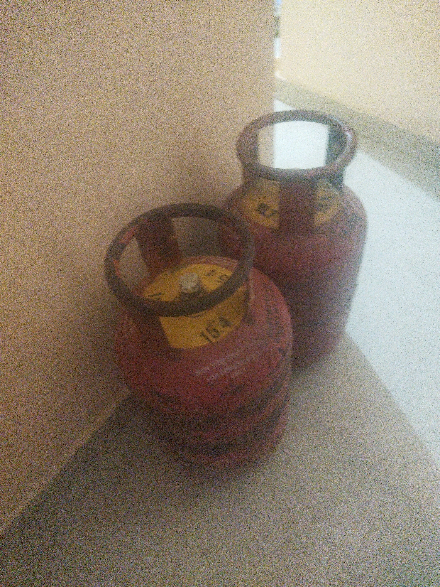 Two red domestic gas cylinders are placed on a beige floor in front of a light beige wall. The cylinder in the foreground is smaller than the one in the background. Both cylinders have a yellow label with black text and numbers. The smaller cylinder has the number "154" clearly visible on its label. The larger cylinder has a label that is partially obscured, but some characters are visible. The overall scene is indoor and appears to be a domestic setting.
