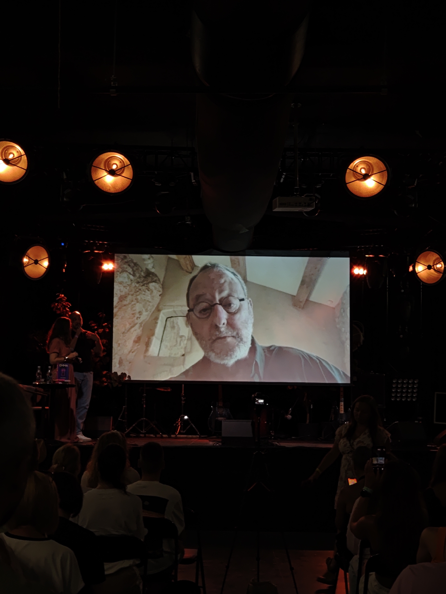 The image captures a dimly lit indoor event, likely a presentation or performance, with a prominent projection screen displaying a close-up of a man's face wearing glasses and a beard. The man appears to be speaking or being recorded. On the stage, a woman in a patterned dress is visible, facing towards the audience, and holding a phone. To the left of the screen, a man and a woman are also on stage, with the man gesturing. The audience members are seated in the foreground, mostly obscured by shadows and darkness, with some holding up their phones. The environment is characteristic of a venue with stage lighting, evident from the spotlights visible in the upper part of the image. The mention of Lviv, Ukraine is not supported by any visual evidence within the image.