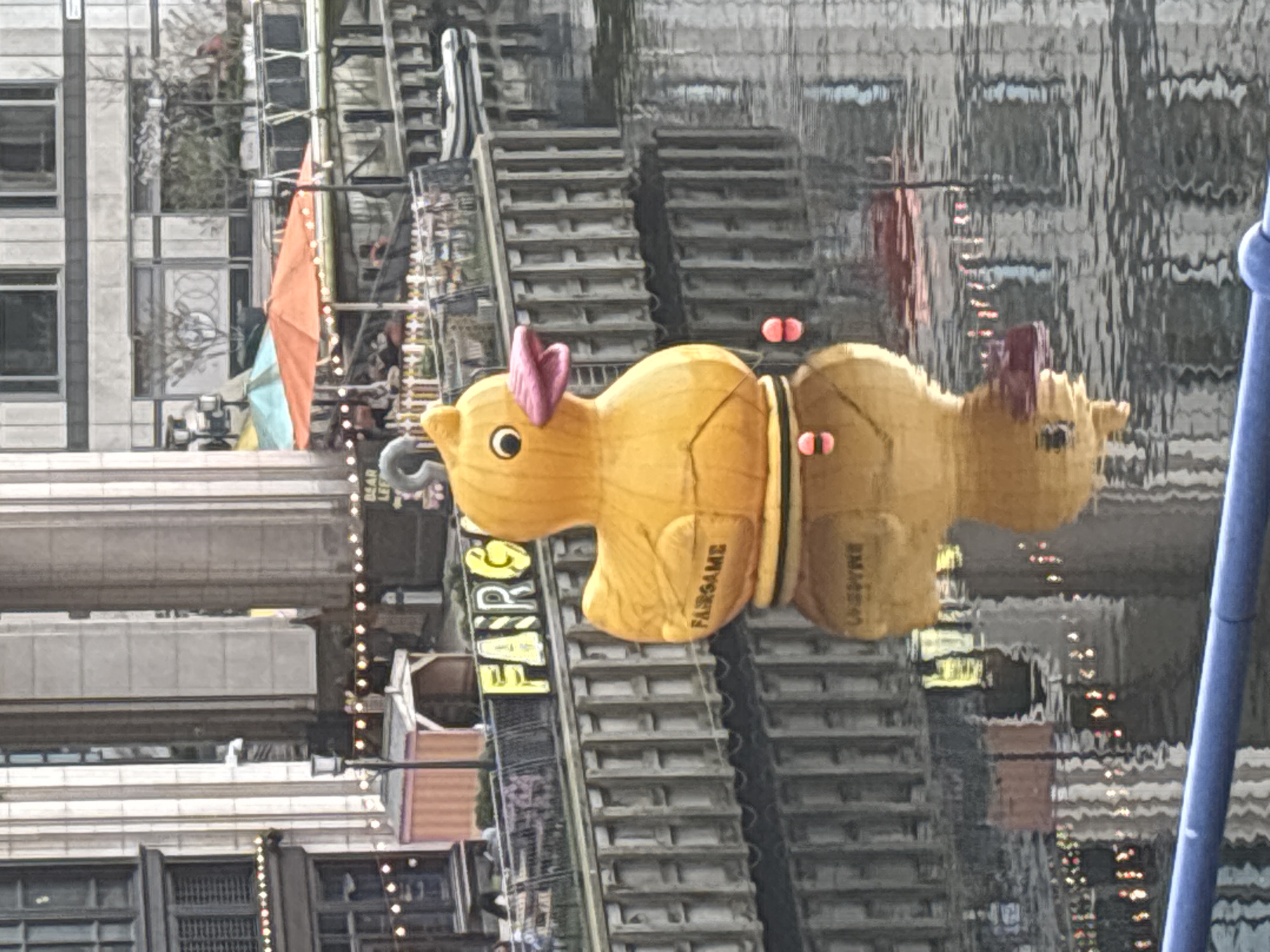 The image shows a large rubber duck floating on water. The reflection of the duck is visible in the water, along with the reflection of buildings. The duck has a pink bow on its head and the words "FAIRGAME" are written on its side. The background features a building and structures similar to stairs, with string lights strung around them. The location is identifiable as London, United Kingdom based on the presence of rubber ducks and building architecture often seen floating on the River Thames as part of festive displays.
