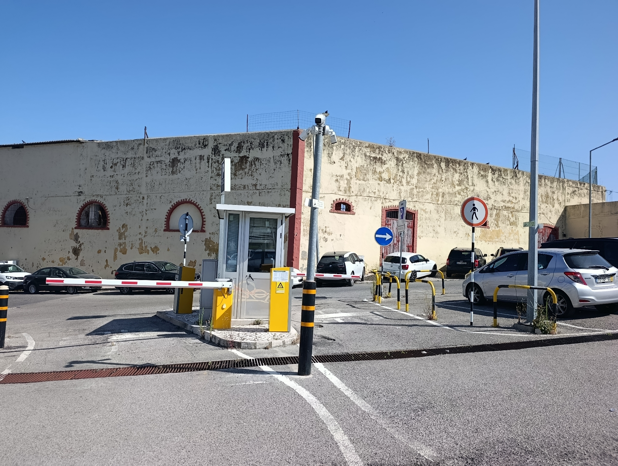 The image shows a parking lot with several cars parked, a security booth, and a barrier gate. A tall pole with a camera is also visible. The building in the background appears to be a warehouse or factory. The sky is clear blue. The scene is likely located in the industrial area of Setúbal, Portugal, near the port, given the type of building and the presence of security measures for a parking lot.