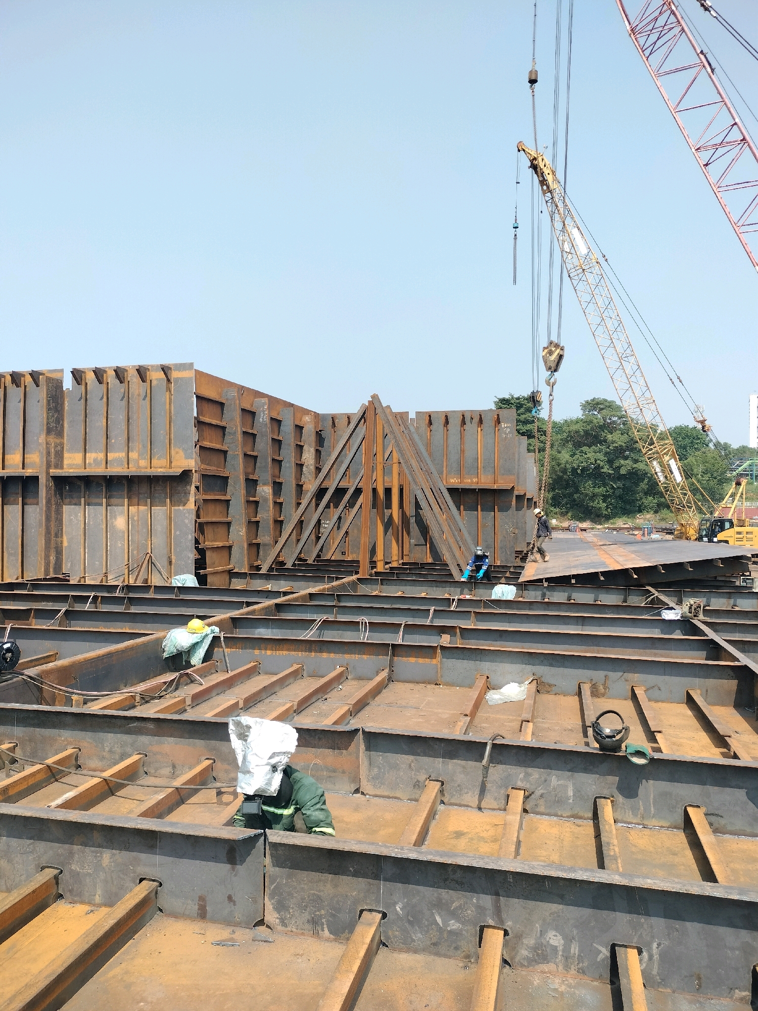 This is a forensic, matter-of-fact description of a construction site in Batam, Indonesia. The image depicts a large-scale metal fabrication or shipbuilding operation. In the foreground, a partially constructed metal structure with numerous steel beams and plates is visible. Several workers in protective gear are present, some appearing to be actively working. One worker in a green jumpsuit and helmet is crouched, possibly welding or inspecting. Another worker in blue gloves and a white hard hat is standing and appears to be directing or observing.
In the mid-ground, massive pre-fabricated steel walls are erected, forming complex angles and sections of a larger structure. These walls have numerous vertical and horizontal supports, indicating their immense size and weight. A yellow crane is positioned in the background, with its boom extended over the construction area, suggesting heavy lifting operations are underway. Another red crane, a lattice boom type, is also visible in the upper right of the frame. The environment appears to be an outdoor industrial area under a clear blue sky. Trees and some distant buildings are visible behind the construction site, suggesting it is located within an industrial zone of Batam. The overall impression is one of active, heavy industrial construction.