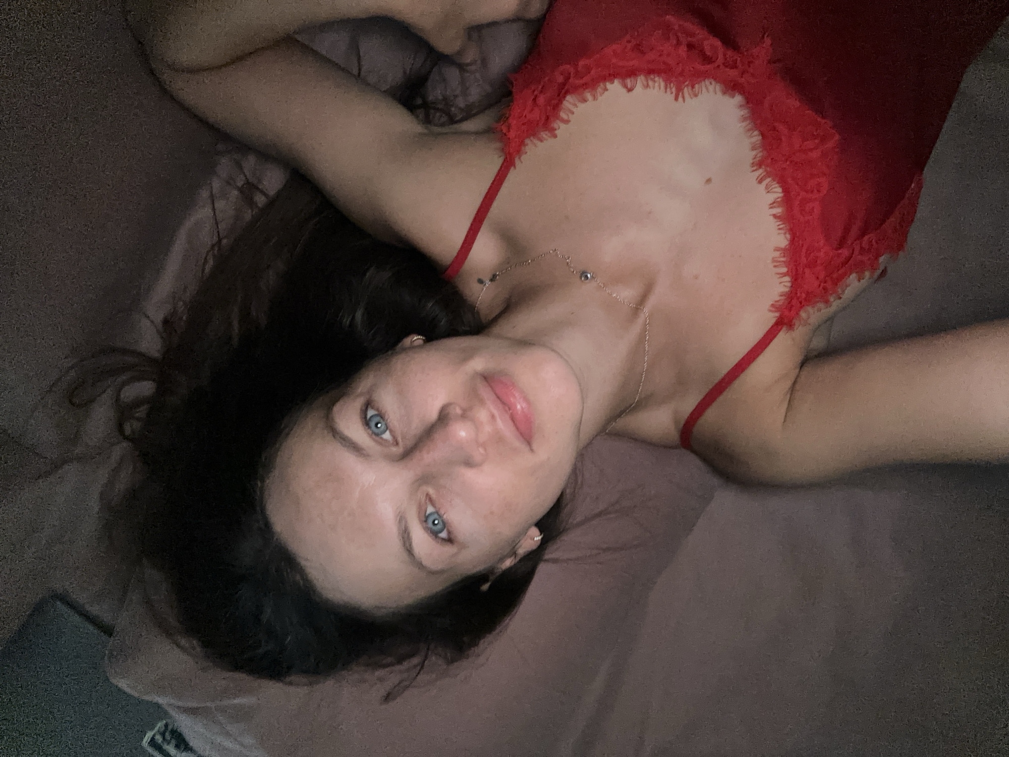 A woman lies on a bed in what appears to be a dimly lit room. She is wearing a red negligee with lace trim. Her hair is dark and spread out on the bedding. She is looking up at the camera with blue eyes. The background is a dark, textured surface, possibly a wall or headboard.