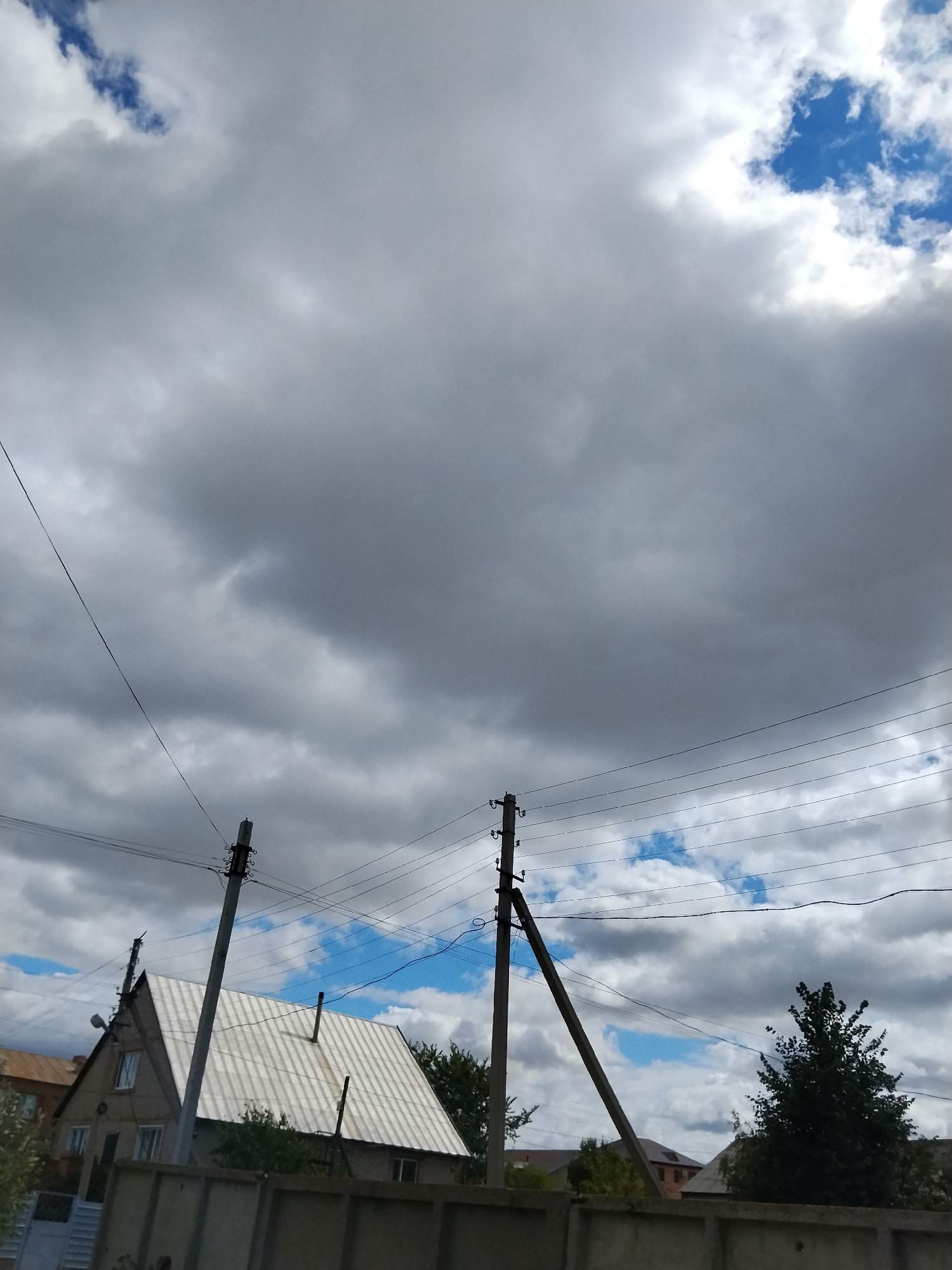 This image captures a cloudy sky over a residential area in Kokshetau, Kazakhstan. In the foreground, a concrete fence runs horizontally. Behind the fence, a house with a light-colored, corrugated metal roof is visible, featuring several windows and a chimney. Two utility poles with electrical wires crisscross the scene, one tall and straight, the other leaning at an angle. A dark green tree stands to the right of the house. The sky is dominated by a mix of dark and light gray clouds, with patches of blue sky showing through in places. The overall impression is one of a somewhat overcast day in a suburban setting.
