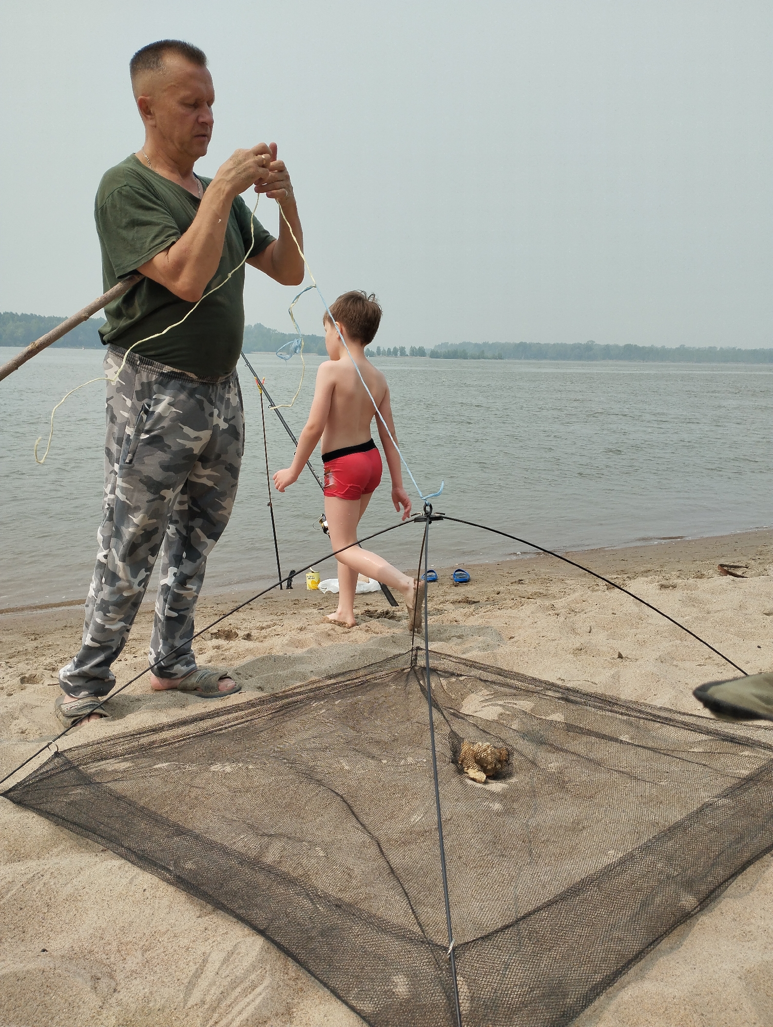 An adult male wearing a green t-shirt and camouflage pants is standing on a sandy beach near a body of water. He is holding a fishing rod and appears to be untangling some fishing line. A young boy, wearing red swim shorts, is walking away from the man towards the water. In the foreground, a fishing net lies open on the sand, with a small object, possibly bait, inside. Fishing rods are visible in the background, along with a distant treeline and a hazy sky. The scene is consistent with a fishing activity on the shore of the Ob River in or near the village of Bibikha, Russia.