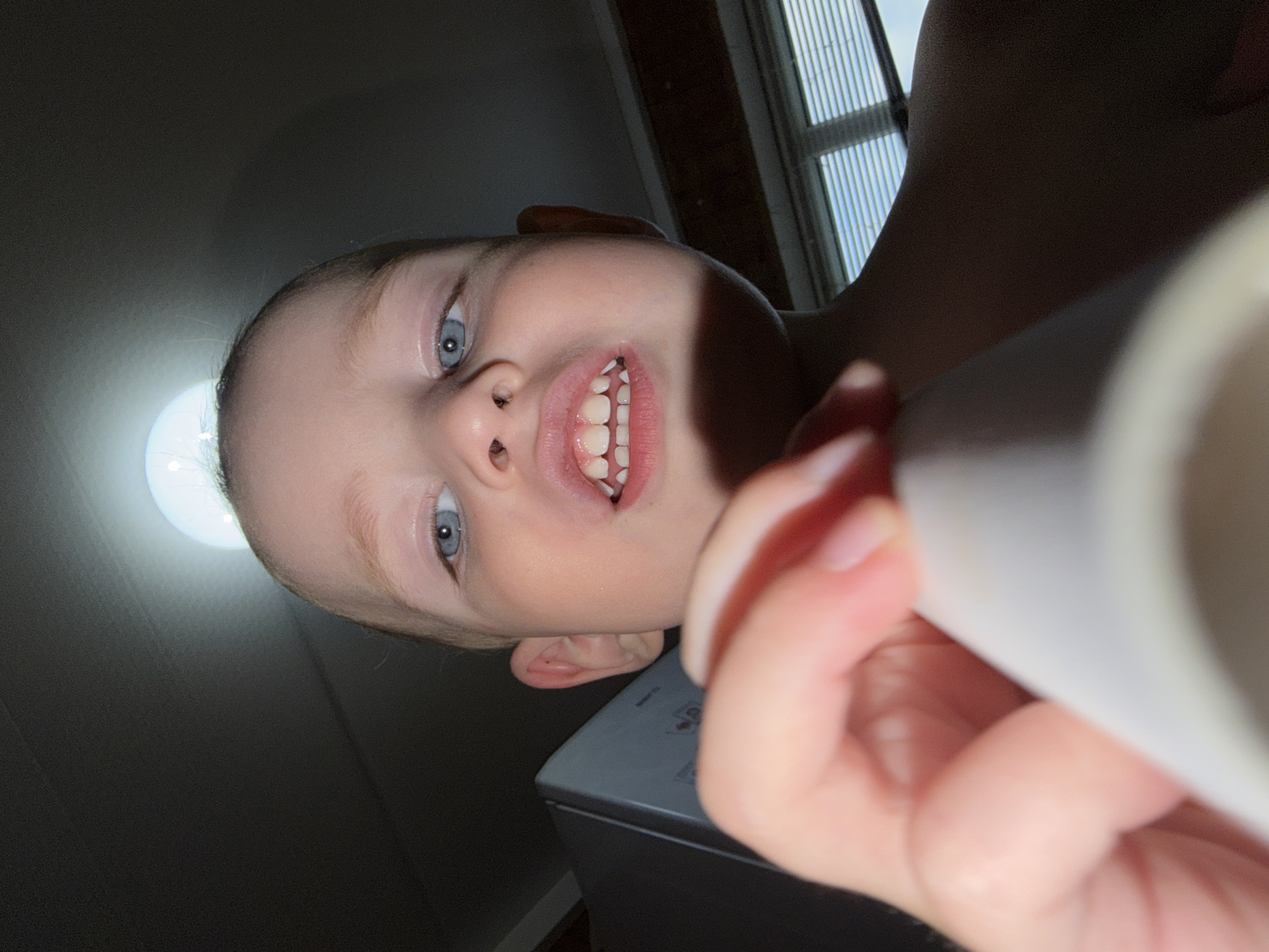 A child with blonde hair and blue eyes is looking up at the camera. The child is holding a white object in their hand. The background shows a window and a light fixture. The location is Уральское, Russia.