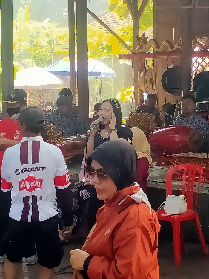 The scene takes place in an outdoor, covered area at a location in Masaran, Indonesia, with coordinates -7.471 latitude and 110.957 longitude. It appears to be a performance space, with multiple people seated, a woman singing into a microphone, and a man playing a traditional Indonesian musical instrument. The woman is wearing a black shirt and black headscarf, while the man is wearing a black shirt with a floral pattern. In the background, several individuals are seated, with some playing traditional instruments. There are several brightly colored, traditional musical instruments in the background, and two chairs with cushions, one with a small white purse on it. The background includes a partially obscured blue umbrella, a green leafy plant, and a wooden structure. The light is bright, indicating a sunny day. The atmosphere is casual and relaxed. The scene captures a snapshot of a cultural performance in Masaran, Indonesia.