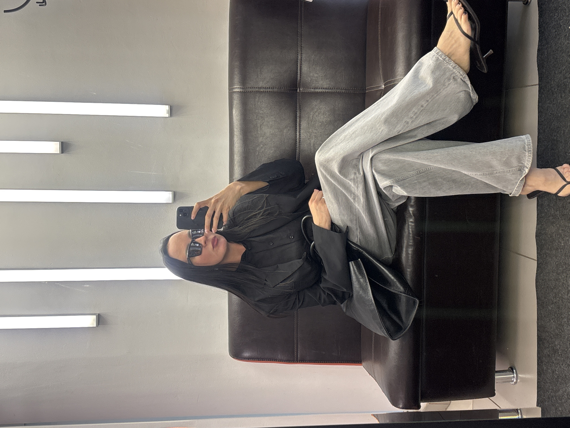 A woman is reclining on a dark brown leather sofa, taking a selfie with her smartphone. She is wearing black sunglasses, a black button-down shirt, light gray jeans, and black heeled sandals. A black leather tote bag sits beside her on the sofa. The background is a light gray wall with several horizontal fluorescent lights. The image was taken indoors. There is no other information that could identify that the location is in Chaikovsky, Russia.