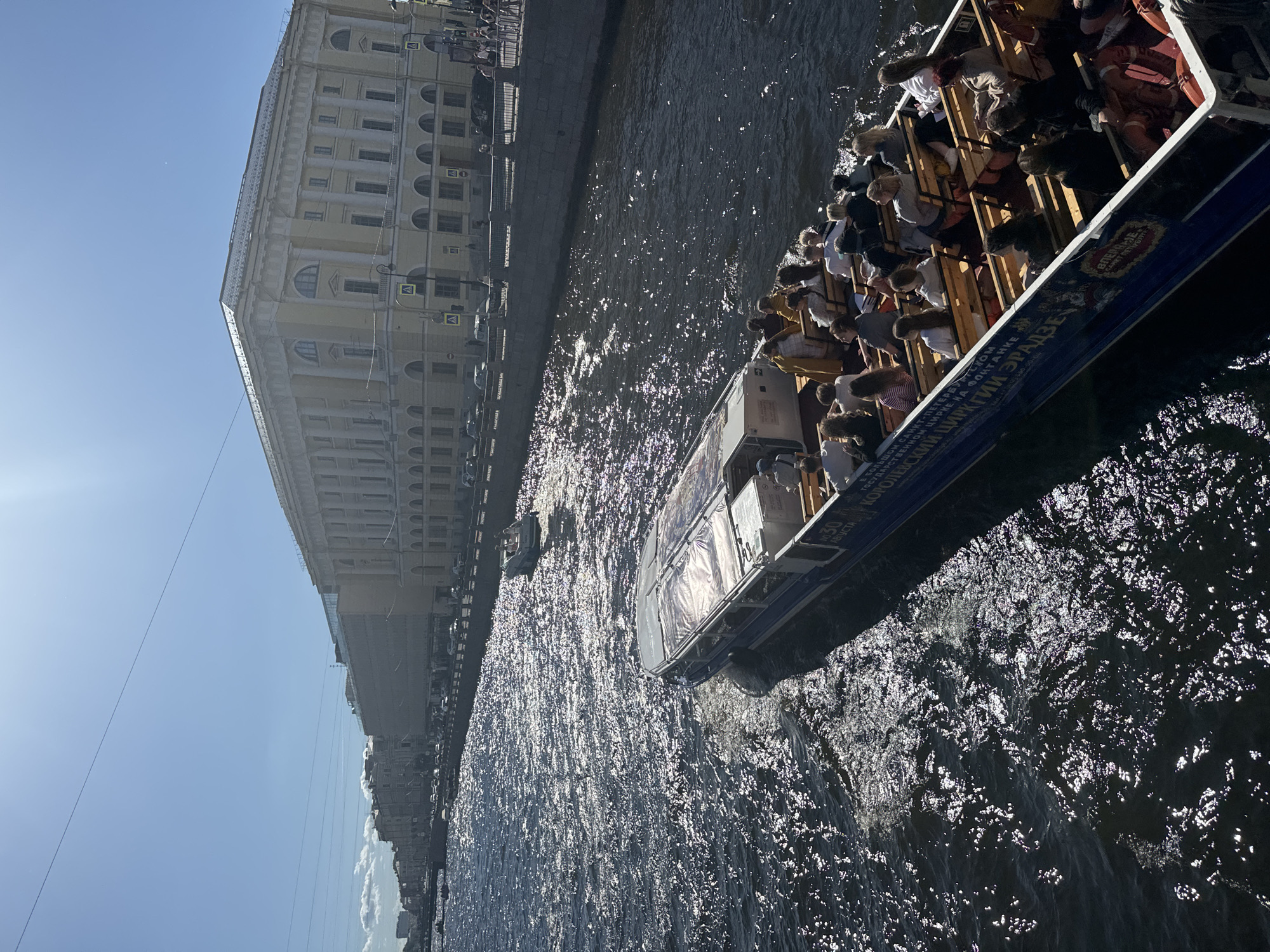 An aerial shot captures a bright, sunny day in Saint Petersburg, Russia, with a prominent tour boat filled with passengers navigating a wide canal. The boat, a long, open-topped vessel with blue and white paint, is moving away from the camera, creating a wake in the dark, reflective water. The passengers, seated on wooden benches, are mostly facing forward, observing the city. Many appear to be tourists, with diverse clothing and hairstyles.
The canal is lined with a stone embankment and a promenade on the right side, where several people are visible standing and walking. Beyond the promenade, a large, ornate yellow building dominates the skyline. This building, characterized by its classical architecture, columns, arched windows, and a flat roof, is situated along the canal. Further down the canal, more buildings of similar architectural style line the waterway, creating a sense of historic urban architecture. The sky is clear and blue with a few wispy clouds. Two smaller boats are also visible in the canal, one further in the distance and another closer to the embankment. The overall scene depicts a leisurely day of sightseeing on the canals of Saint Petersburg.