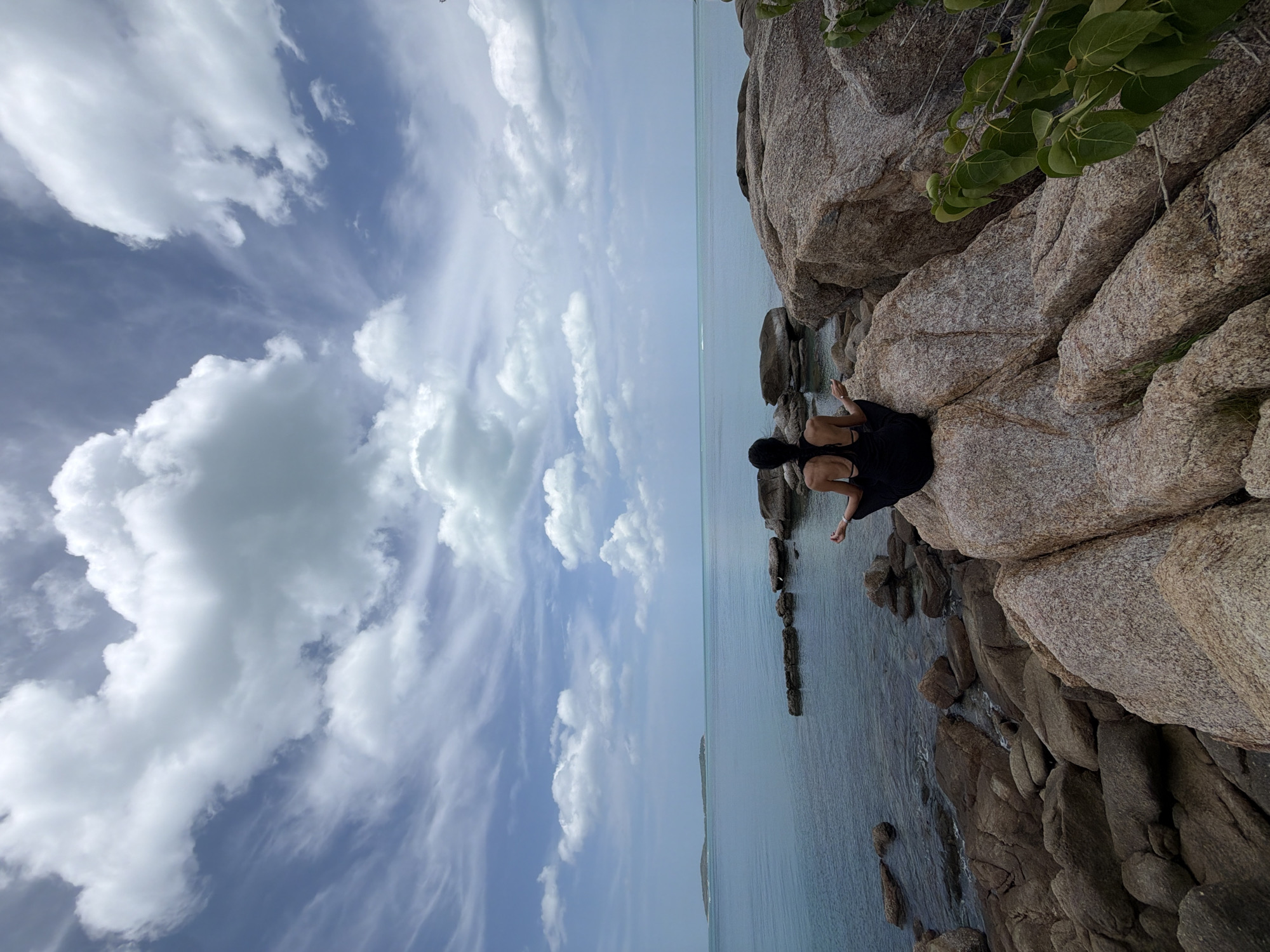 A person in a black dress meditates on a rock formation by the sea in Ko Samui District, Thailand. The individual is seated with legs crossed and hands resting on their knees, facing the ocean. The sea is calm with a few scattered rocks visible in the water. In the distance, a small island is visible on the horizon. The sky is partly cloudy with a mix of white cumulus clouds and blue sky. The foreground features large, rough, beige-colored rocks. Green foliage is present on the right side of the image.