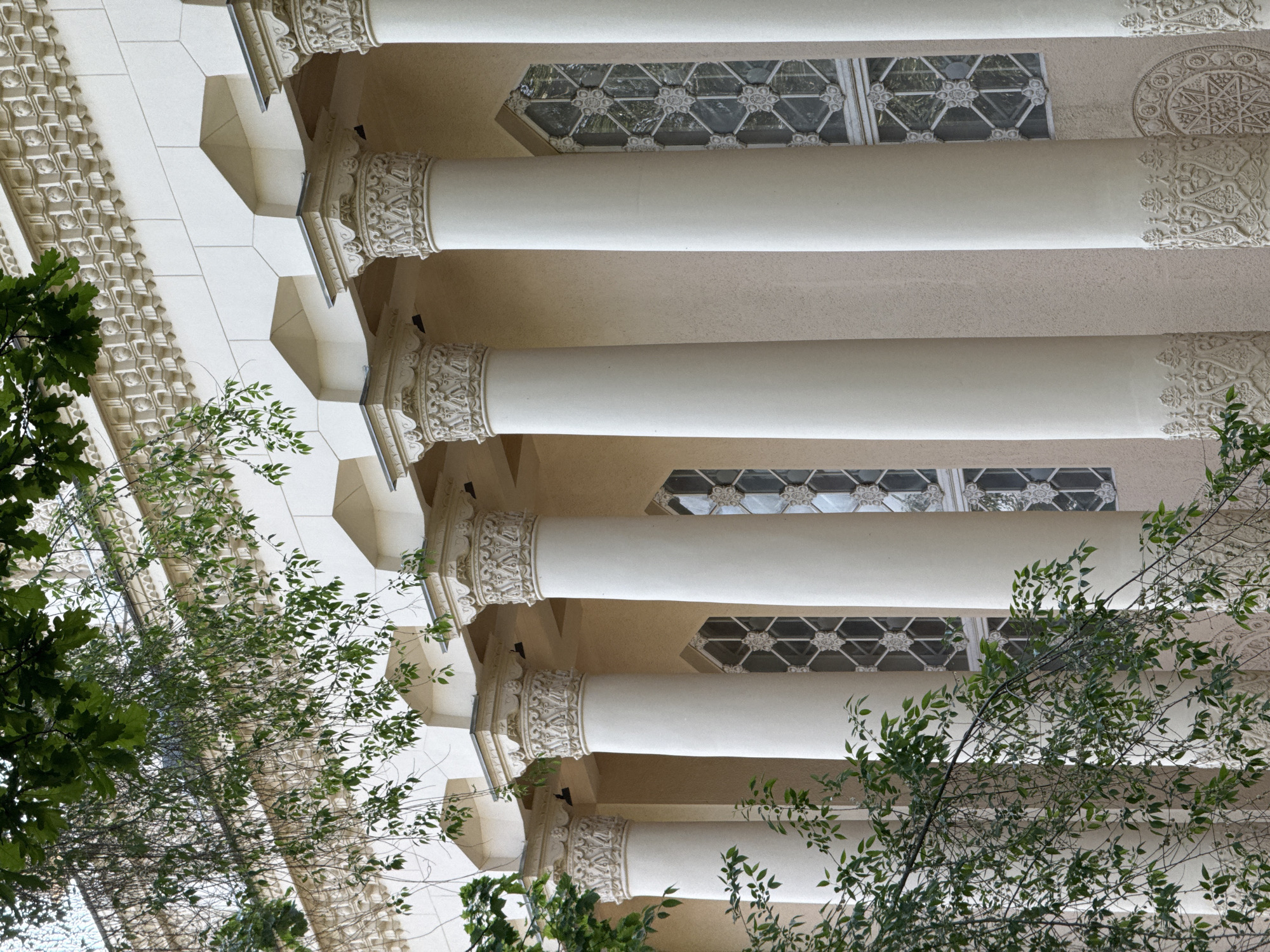 The image shows a low-angle view of architectural elements, possibly exterior pillars, with ornate decorations. The location is likely Moscow, Russia, based on the architectural style, which includes white pillars, patterned surfaces, and window details. Greenery from trees is visible in the foreground, partially obscuring the view of the structure. The scene appears to be taken during daylight hours.