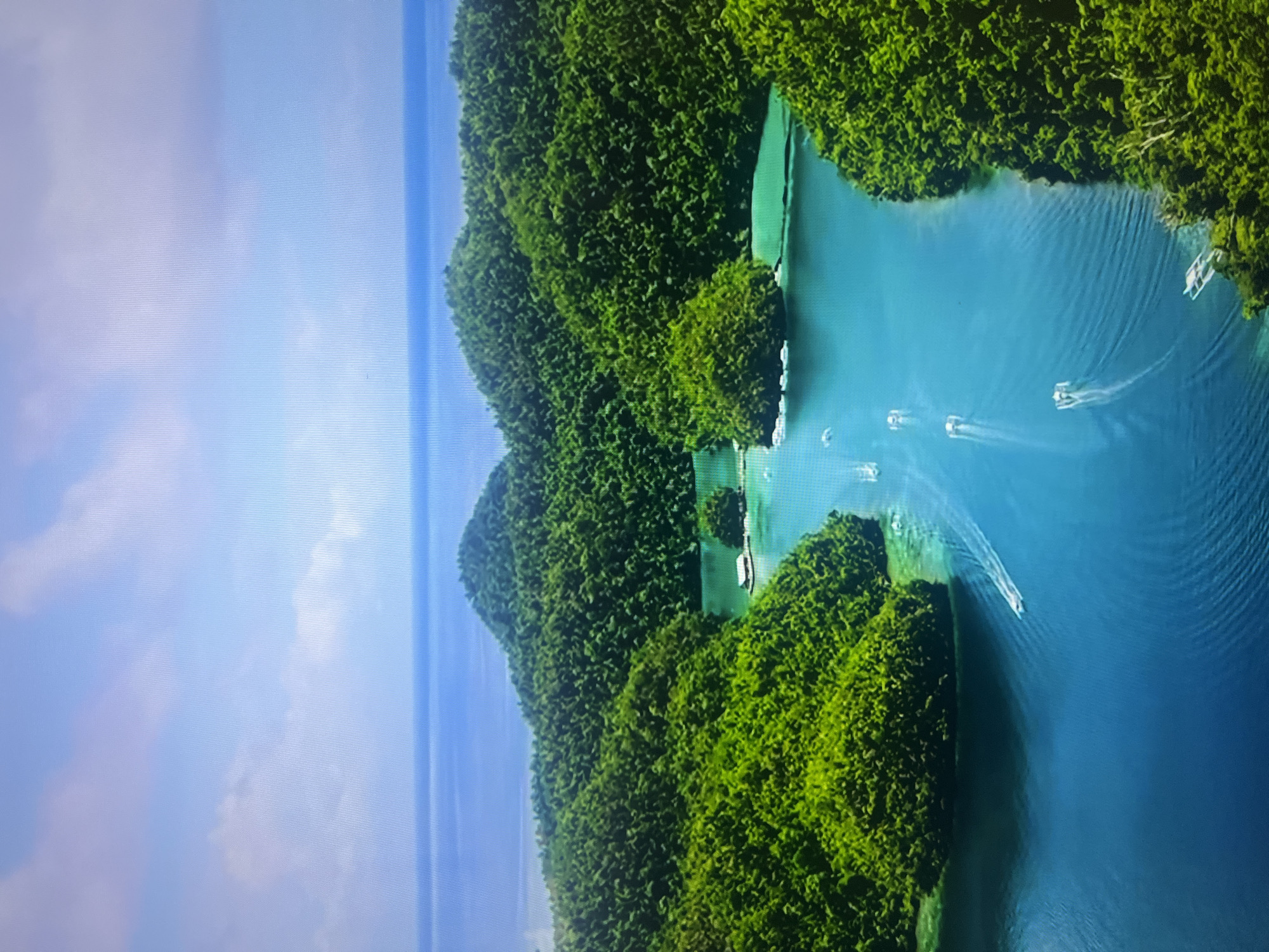An aerial view showcases a serene tropical paradise with lush green islands dotting a vibrant turquoise ocean. The islands are densely covered with dense green foliage, creating a dramatic contrast with the clear blue water. Several boats navigate the calm waters, leaving white trails as they speed across the surface. In the distance, the horizon meets a clear blue sky dotted with scattered white clouds. The scene evokes a sense of tranquility and natural beauty, characteristic of a secluded tropical destination.