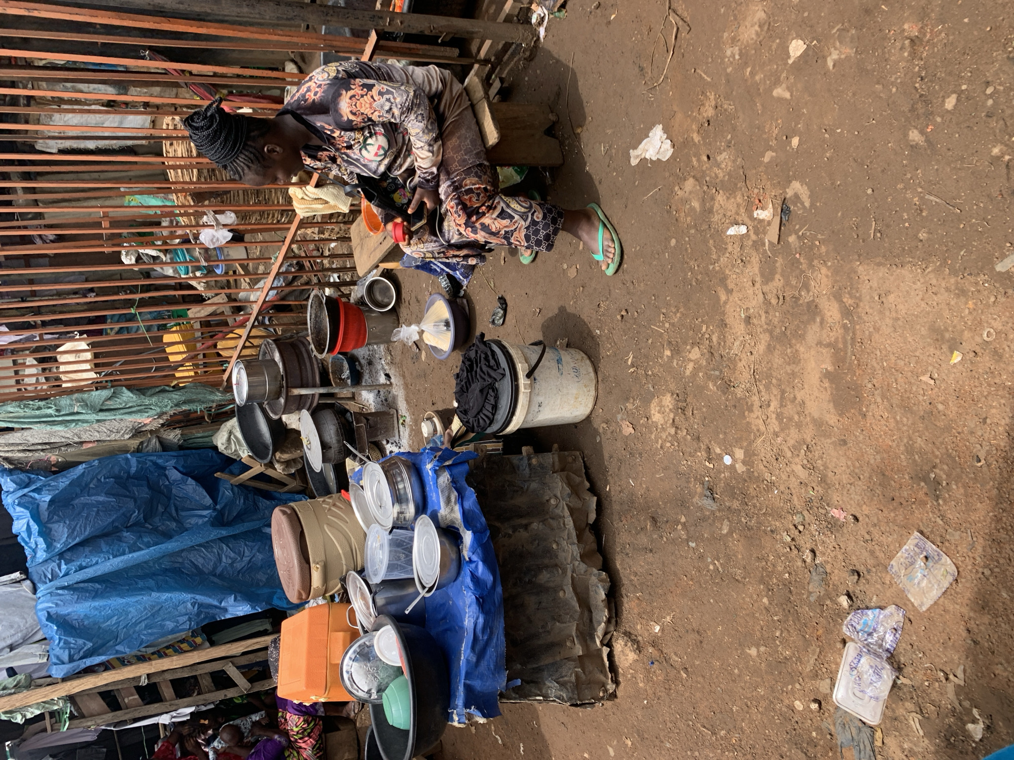 An Ilorin, Nigeria market scene captures a seated Black woman looking at her mobile phone; she's dressed in patterned pants and a matching top and is wearing green flip-flops. A jumble of cookware including pots, pans, and buckets surround her, with some covered by a blue tarp. A makeshift metal structure is above her, and the ground is dirt, scattered with debris.
