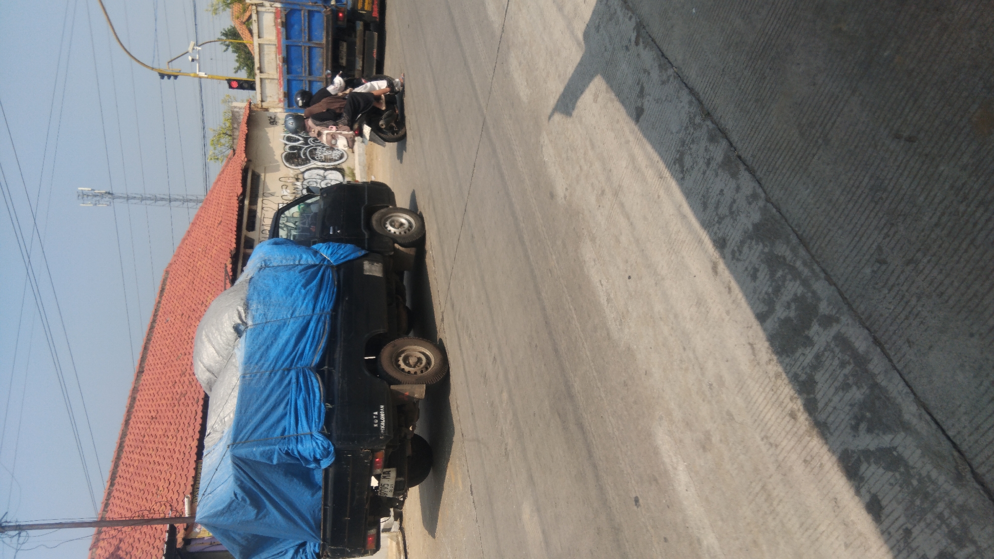 In the city of Batang, Indonesia, on a bright, sunny day, a black pickup truck with a blue tarp covering its cargo is parked on a road. The truck has a license plate that reads "B 19795 MA". Behind the pickup truck, a blue flatbed truck is also visible. Two people are riding on a motorcycle, with one person driving and the other as a passenger. The background features buildings with red tile roofs and some graffiti on a wall. A traffic light is visible, indicating a possible intersection. The road surface is concrete with visible tire marks and a large shadow cast by an unseen object.