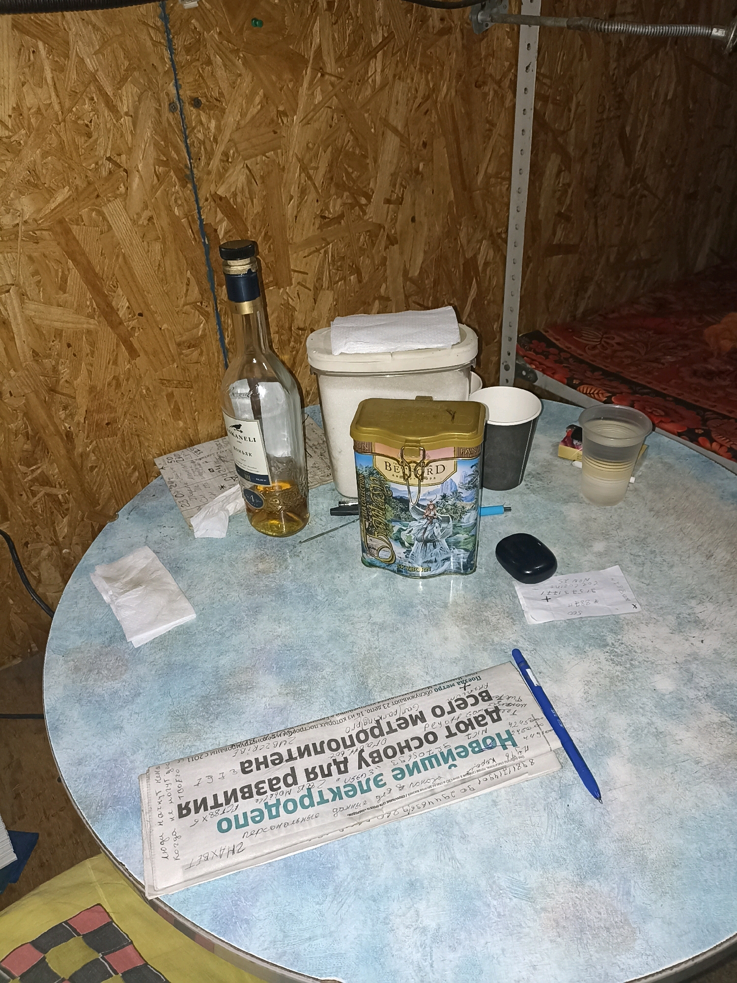 The image depicts an indoor scene, likely a room with unfinished wooden walls. On a light blue circular table, several objects are arranged. To the left, a clear glass bottle with a dark blue cap contains a golden liquid, possibly alcohol. Next to it is a white plastic container with a white lid, and a stack of white paper napkins.
In the center of the table, a gold-colored tin with an illustration of a woman and a waterfall is visible. In front of the tin is a black, oval-shaped electronic device. To the right of the tin, a grey paper cup and a transparent plastic cup filled with a light-colored liquid are placed.
A folded newspaper lies on the table, with its headline visible and some text. A blue pen rests on top of the newspaper. Another piece of paper with handwritten text and numbers is situated to the right of the black electronic device.
In the background, a metal shelving unit is partially visible, and beyond that, a bed with a patterned red and black textile is present. The overall impression is one of a casual, possibly temporary, living or working space.