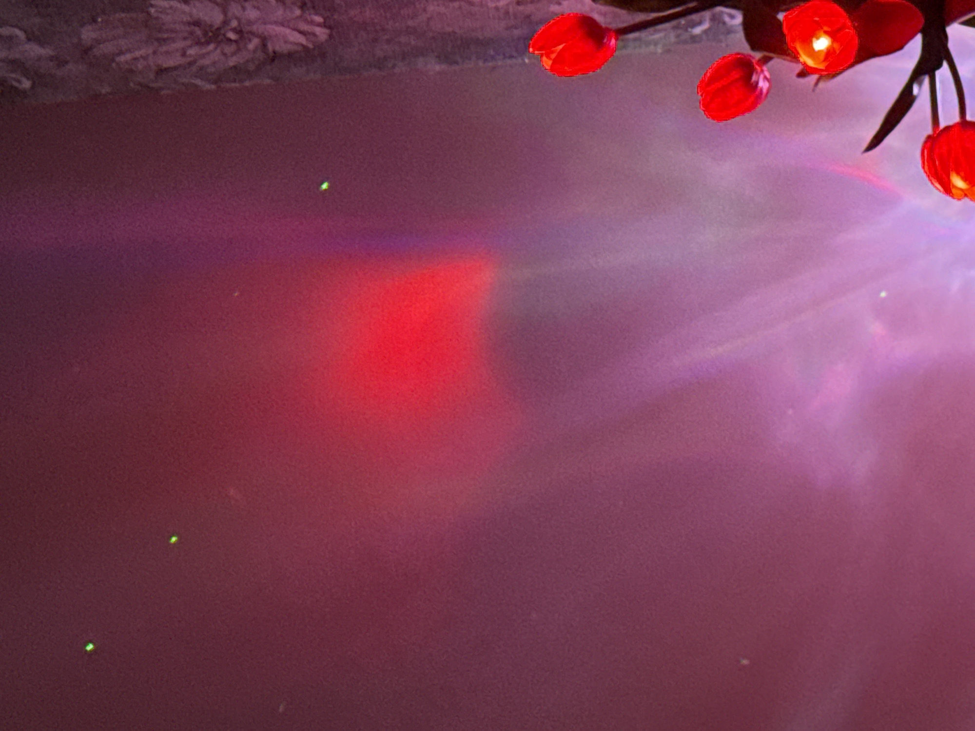 A low-angle shot reveals red, tulip-shaped lights illuminating a blurred, violet-tinged background. The top of the frame features a textured, greyish surface, possibly a ceiling. The majority of the scene is a smooth gradient transitioning from deep red to lighter violet, with a few small green dots scattered throughout, possibly reflections or defects. The light from the tulips spreads out, creating a soft, diffused glow around them. There is no indication of people, specific objects besides the lights, nor of a location in a city.