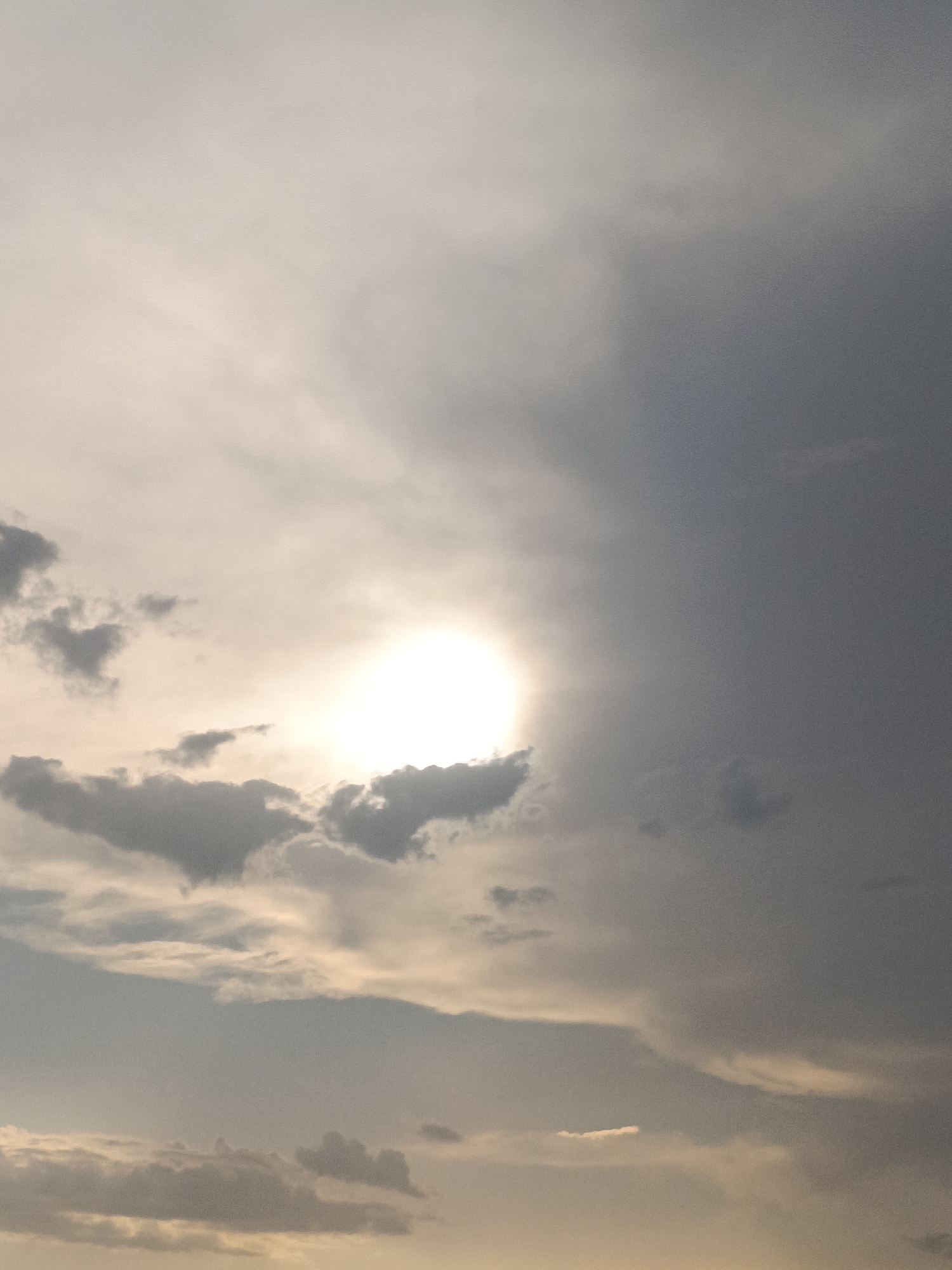 The image depicts a sky filled with a variety of clouds. The sun is visible, partially obscured by clouds, casting a bright, diffused light. The clouds range in color from bright white where illuminated by the sun, to various shades of gray, with some appearing darker and more ominous. There is a sense of depth in the cloud formations. The overall lighting suggests either sunrise or sunset. The image was captured in Cabanes, Spain. There are no people or discernible man-made objects in the frame, only the sky and clouds.