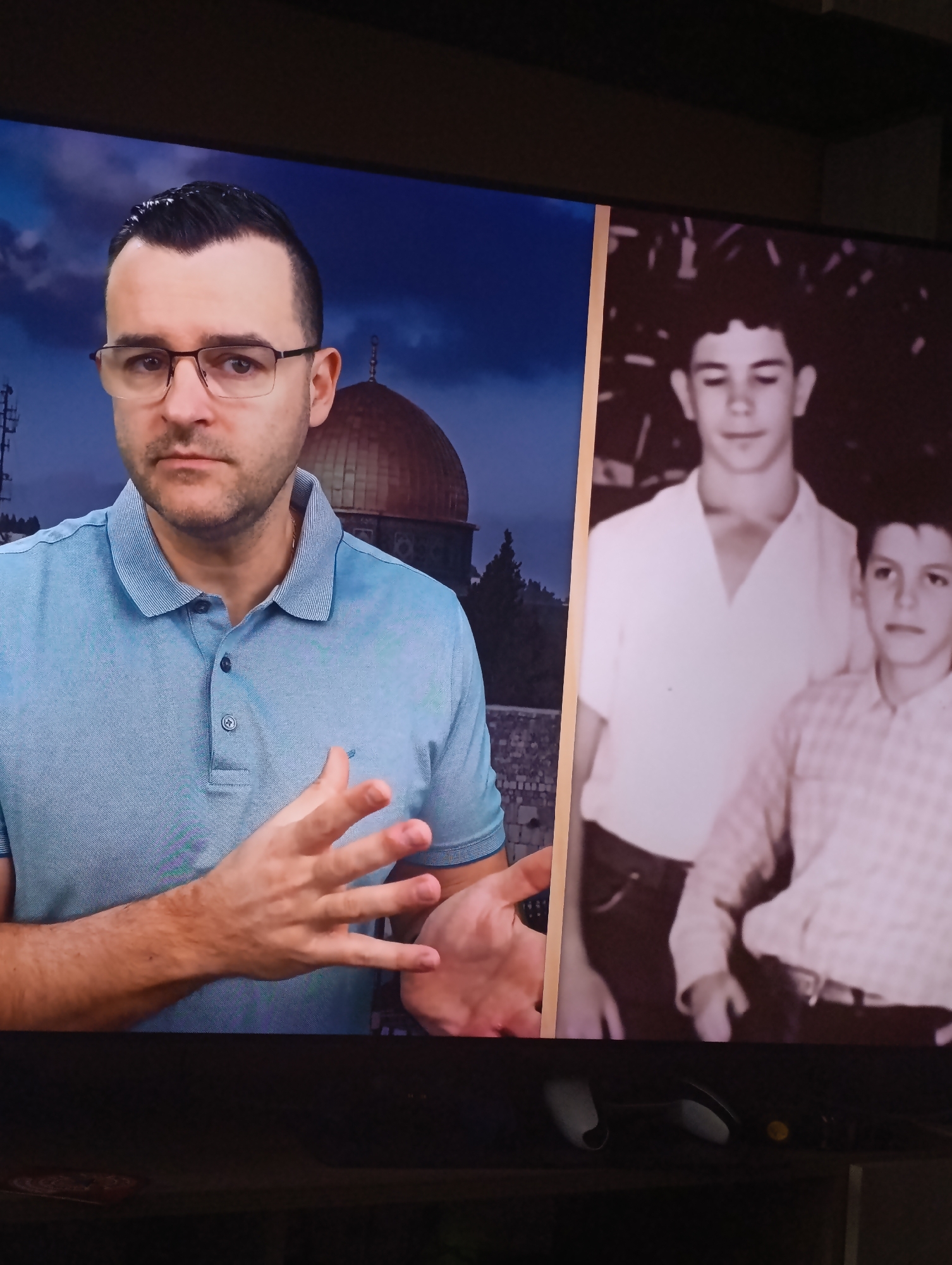 The image shows a man in a light blue polo shirt and glasses standing in front of a backdrop that appears to be the Dome of the Rock in Jerusalem. To the right of the man is a black and white photograph of two young boys. The photograph is held up to the camera. The location in Plovdiv, Bulgaria, is not apparent in the image.