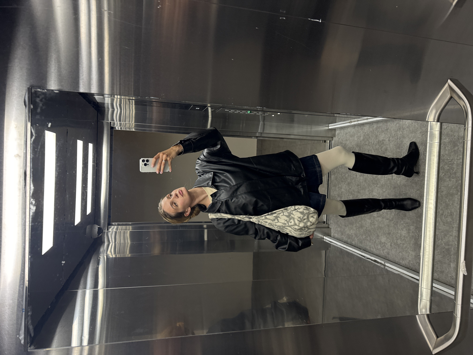 A woman stands in an elevator in Unknown, Unknown, taking a selfie with a white smartphone. She is wearing a black leather jacket, a dark skirt, white tights, and black knee-high boots. She has a patterned handbag slung over her shoulder. The elevator has a stainless steel interior with vertical lines on the walls and a mirrored back panel.