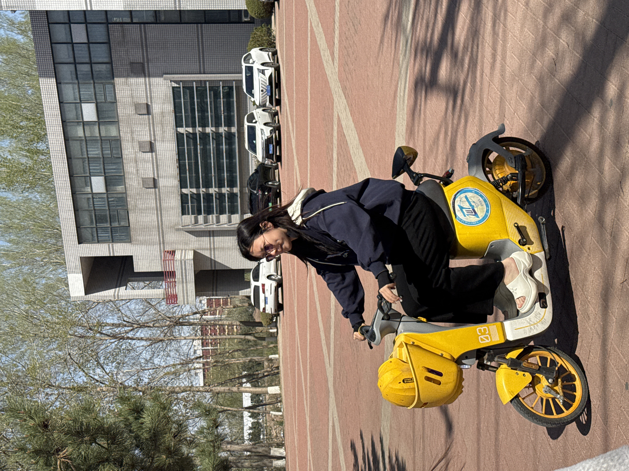 A medium shot depicts a person seated on a yellow scooter parked on a reddish-brown paved surface. The person is facing to the left, wearing a navy blue jacket and black pants. The scooter has a visible "03" marking on the side. Behind the person and scooter, a modern building with numerous windows is visible, along with parked cars. Trees are present in the background to the left. The scene is likely located in Zhongtun, China, based on the prompt. The image is rotated 90 degrees counterclockwise.