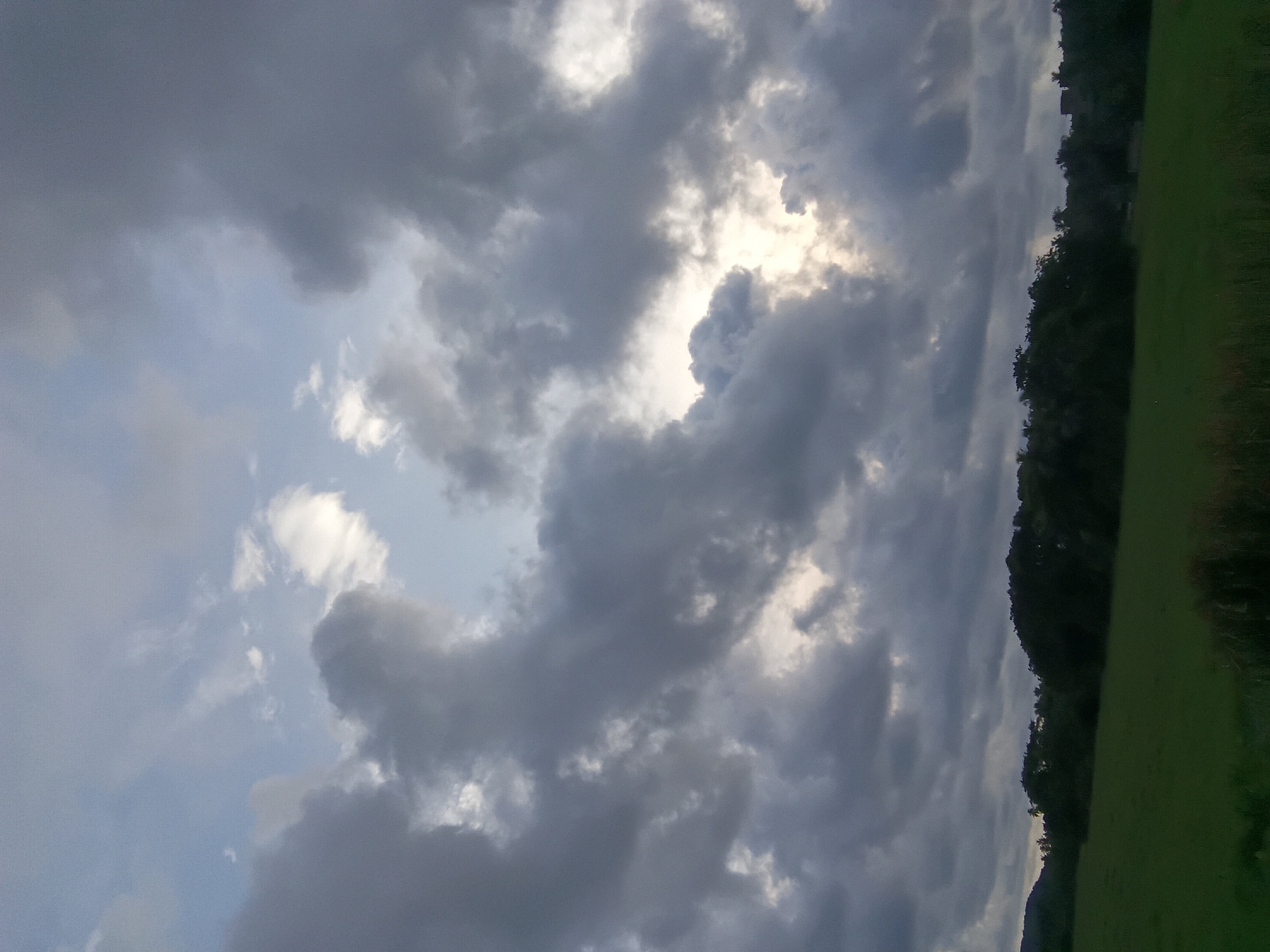 The image depicts a cloudy sky with a break in the clouds revealing sunlight. On the right side of the frame, there's a line of trees and a grassy field. The photograph appears to have been taken outdoors, possibly in the vicinity of Sodiem, India, though specific landmarks cannot be discerned from the image alone. There are no people or obvious objects besides the natural elements. The main interaction is between the sunlight and the cloud cover. The environment appears to be natural.