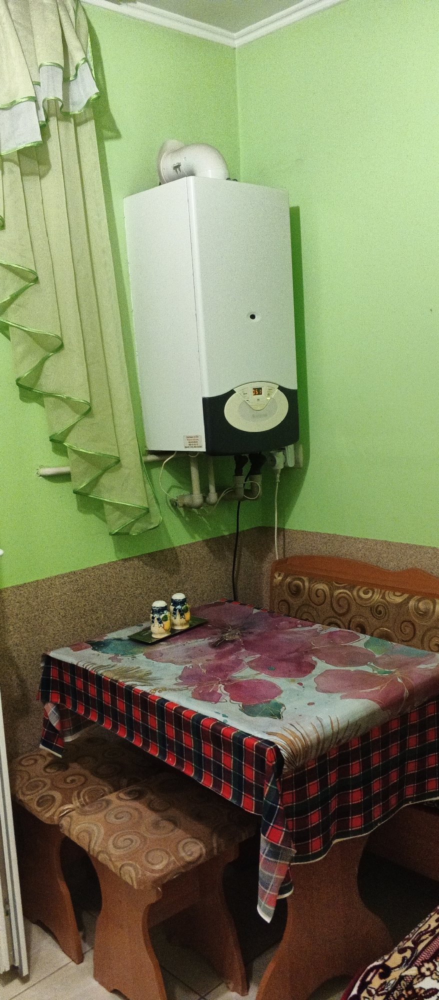 The image depicts an indoor scene, likely a kitchen or dining area, with bright green walls. A white gas boiler is mounted high on the wall, with a white pipe extending from it. In the foreground, a small table is covered with a floral-patterned tablecloth overlaid with a red and black plaid fabric. Two wooden stools with patterned cushions are placed under the table. On the table, there are two salt and pepper shakers on a small tray. To the right of the table, a portion of a bench with a patterned upholstery is visible. A light green curtain with ruffled edges hangs on the left side of the frame. The overall impression is of a functional, lived-in space. Based on the available information, this scene is located in Berehove, Ukraine.