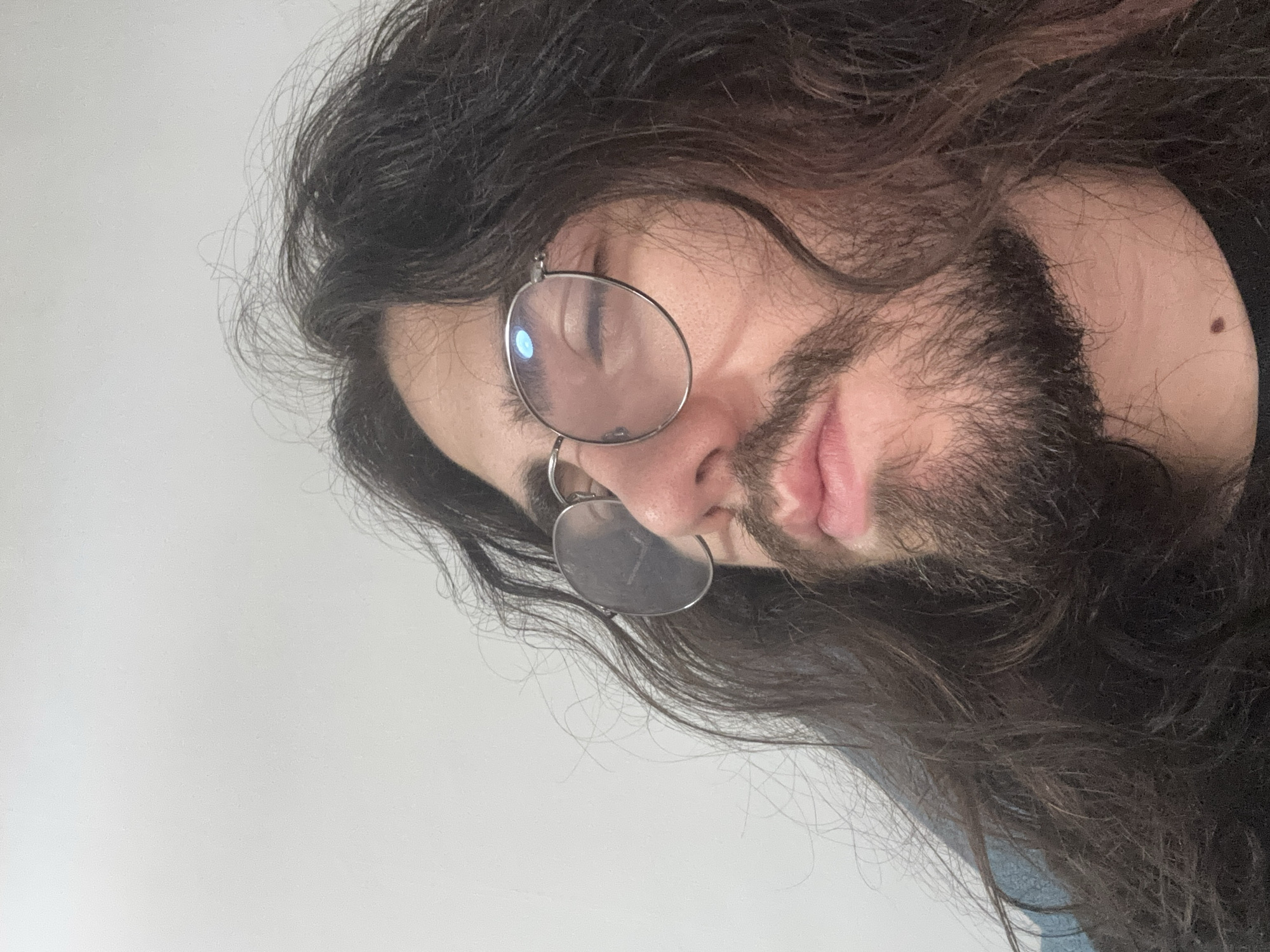 An eye-level, medium shot captures a person's face from the nose up, against a plain white background. The individual has long, dark, wavy hair that partially obscures their face and falls over their shoulders. They are wearing round, metal-rimmed glasses with blue-tinted lenses. Their eyes are closed or nearly closed, and their head is tilted slightly upwards and to the left. A mustache and beard are visible, along with slightly parted lips. The lighting is soft, with a noticeable blue reflection on the left lens of the glasses.