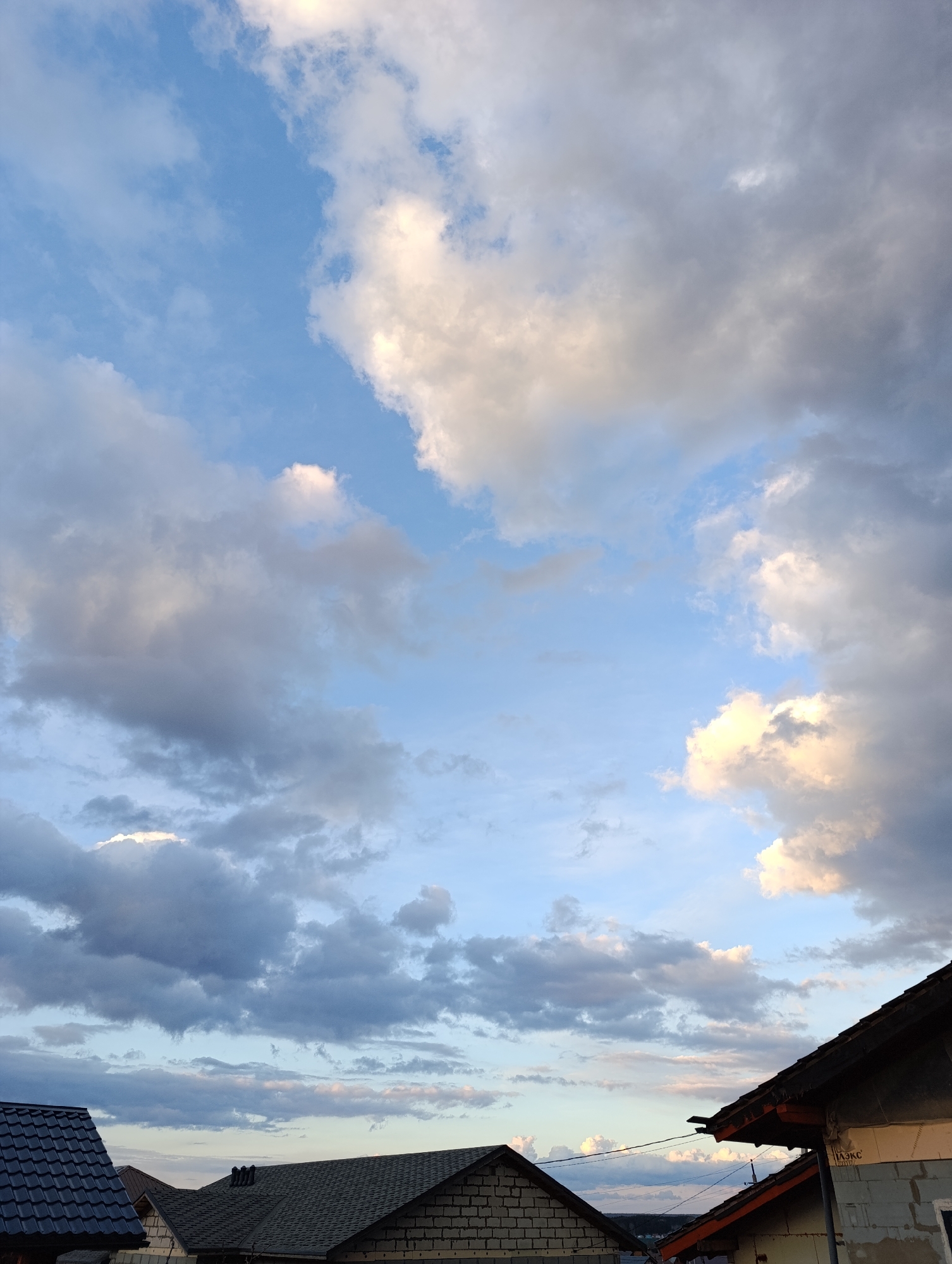 The image displays a vast, dramatic sky dominated by an array of clouds and glimpses of residential rooftops below. The sky features clear patches of bright blue, particularly in the upper left and central areas, contrasting with extensive cloud formations. These clouds vary in color, ranging from bright white and light grey to darker, denser grey masses. Some of the clouds, especially on the right side and near the top, are illuminated with warm golden and soft pinkish hues, indicative of either late afternoon or early evening light.
At the bottom of the frame, parts of several building roofs are visible, suggesting a residential setting. On the far left, a dark, possibly tiled roof is prominent. Towards the center and right, other dark, shingled roofs are seen, along with the upper sections of buildings under construction or recently built, featuring exposed brick or concrete block walls. On the rightmost building, a white label or sign with the Cyrillic word "ЛЭКС" is discernible. No people or specific activities are visible within the frame. The scene conveys a sense of expansive natural beauty combined with human development, set under a dynamic sky.