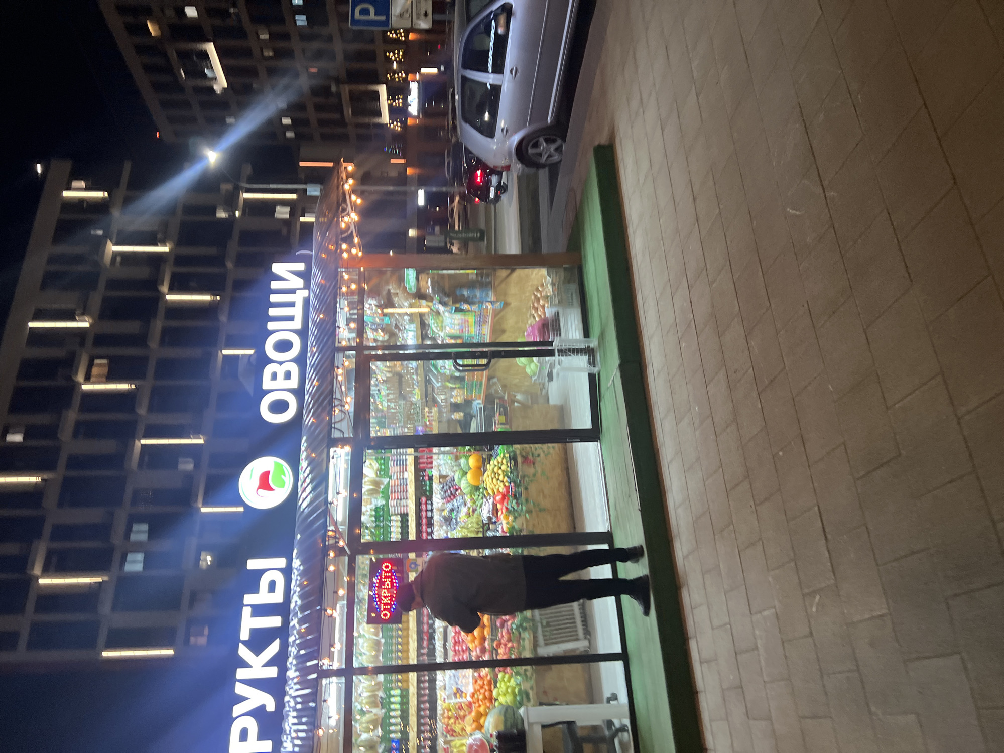 The image shows a street scene in Tyumen, Russia at night. A fruit and vegetable shop ("ФРУКТЫ ОВОЩИ") is visible, illuminated by bright lights and a string of decorative bulbs. A customer is inside the shop, browsing the produce. A car is parked on the street in front of the shop. A modern apartment building is in the background. The sidewalk is paved with rectangular tiles.