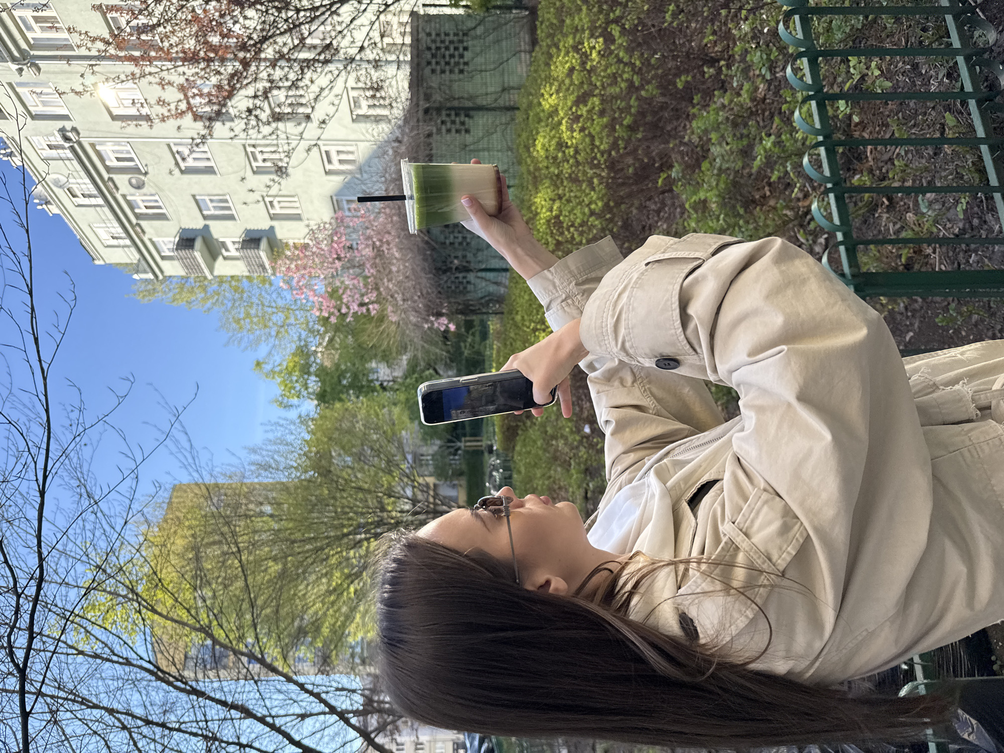 A young woman is depicted outdoors on a bright, clear day, likely in a park or urban green space in Warsaw, Poland. She has long, dark hair, wears glasses, and is dressed in a light-colored (beige or khaki) jacket or trench coat. In her right hand, she holds a smartphone, appearing to take a photograph or video, while her left hand holds up a clear plastic cup containing a layered drink—green on top and an off-white or cream color at the bottom, with a black straw.
The background reveals a multi-story building with a light-colored (pale green or grey) facade and numerous windows, characteristic of urban European architecture. The scene is lush with early spring foliage; some trees have bare branches, others display fresh light green leaves, and a few are adorned with delicate pink blossoms. A vibrant clear blue sky is visible overhead. A dark green metal fence or railing is partially visible in the foreground, bordering the green space. The bright sunlight indicates it is daytime, suggesting a pleasant morning or afternoon. The woman's activity implies she is enjoying the pleasant weather and capturing the scenic environment. No discernible text is visible on the phone screen or the drink cup.