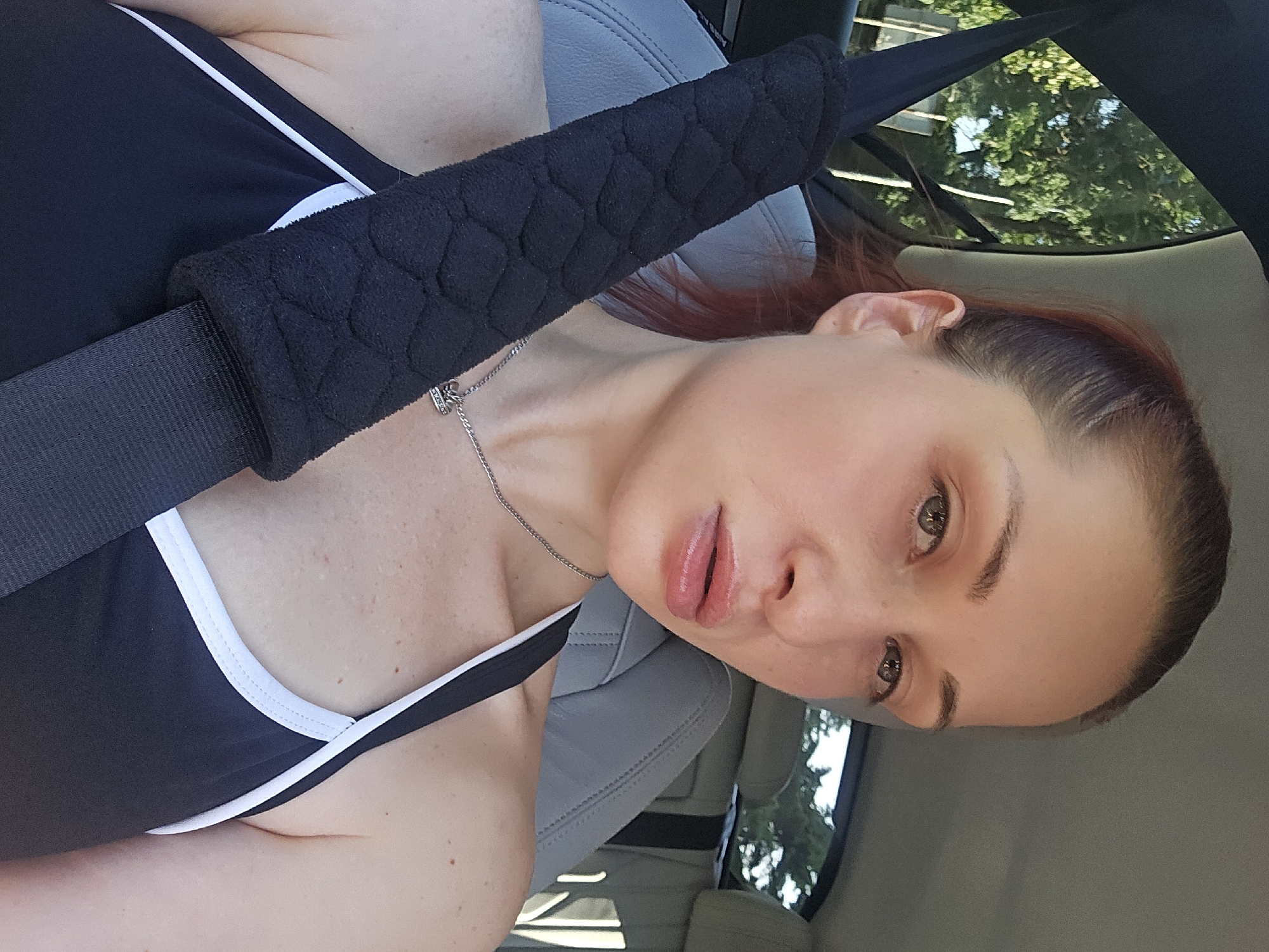 A young woman with reddish-brown hair is taking a selfie from the passenger seat of a car. She is wearing a black tank top with white trim and a silver necklace. A black seatbelt pad is visible across her chest. The interior of the car is gray leather. Through the window, lush green trees are visible, suggesting it is daytime and likely warm weather. The woman's facial expression appears neutral. The scene is set in Burgas, Bulgaria.