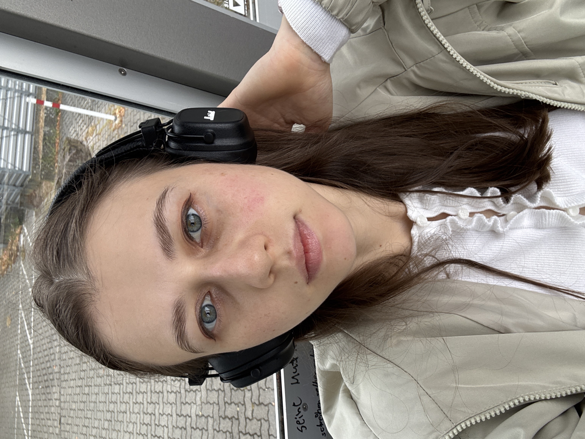 A young woman with long brown hair and blue eyes, wearing black headphones, a cream-colored jacket, and a white ruffled top, is taking a selfie outdoors. She is touching her headphones with her right hand. The background shows a paved area with white lines, a metal fence, and some trees with fallen leaves, suggesting an outdoor urban or semi-urban environment. Text is visible on a metal surface behind her, but it is partially obscured. The scene appears to be in Erlenbach am Main, Germany, based on the provided context.