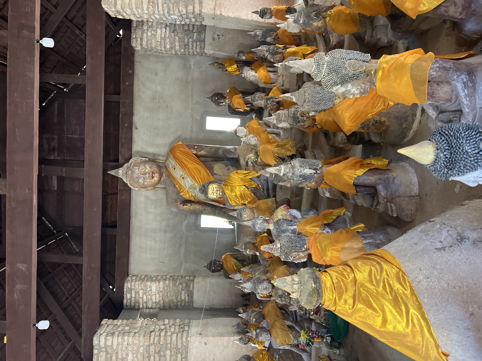 The image depicts a row of Buddha statues, each draped with a yellow cloth, inside a structure with a wooden ceiling and brick or stone walls. The setting is possibly a temple or historical site, and the location is Baan Harn, Thailand. The statues appear to be lined up against the wall, with a larger, more prominent Buddha statue at the forefront. There are a number of smaller statues lined behind it. The statues vary in appearance and condition, showing possible signs of aging or wear. The light source appears to be coming from the right, illuminating the right side of the statues more than the left.