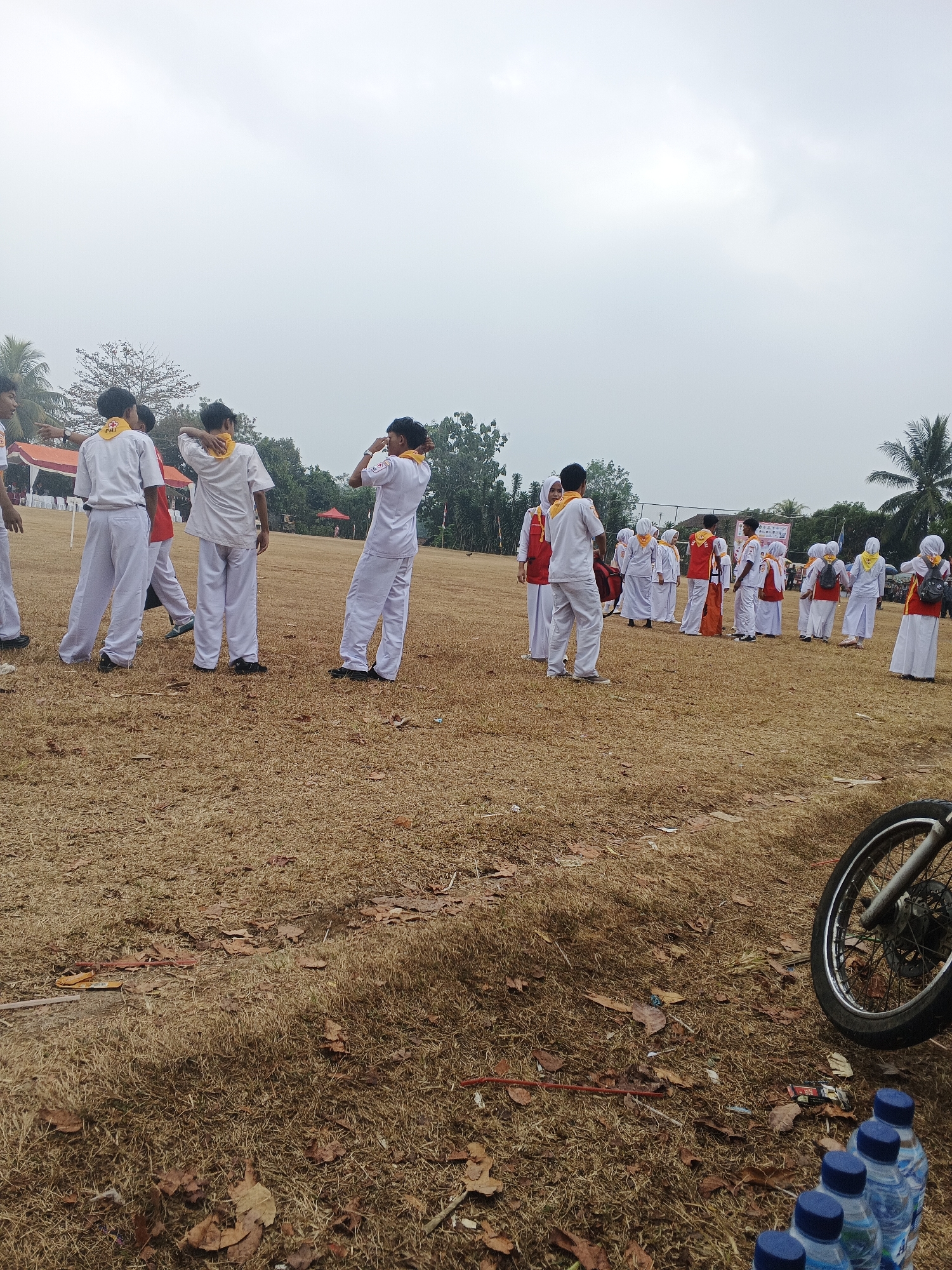 A group of youths in white uniforms and yellow scarves stand on a dry, grassy field. Some are facing away from the camera, touching their necks or heads. In the background, other similarly dressed youths are gathered, some wearing red vests over their uniforms. A few tents and trees are visible in the distance. The sky is overcast. In the foreground, a motorcycle wheel and several bottles of water are on the ground. The scene appears to be outdoors, possibly at a school event or gathering in Warungkiara, Indonesia.