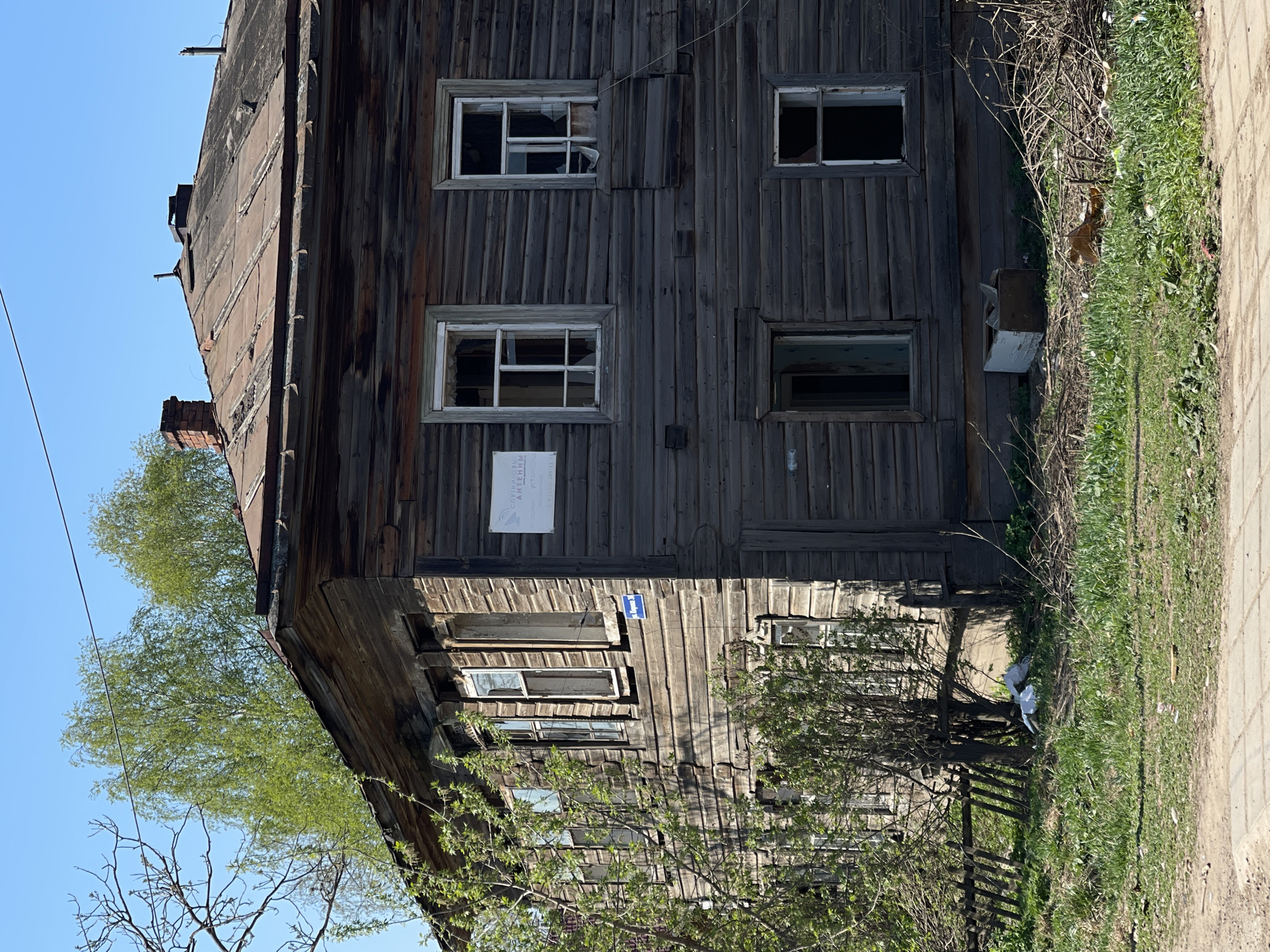 The image shows an old, two-story wooden building in Фоки, Russia. The building appears dilapidated with weathered wood siding, broken windows, and a rusting roof. A sign is attached to the building's exterior. Lush green grass and foliage surround the building's base. A sidewalk runs along the building's right side. The sky is clear and blue.