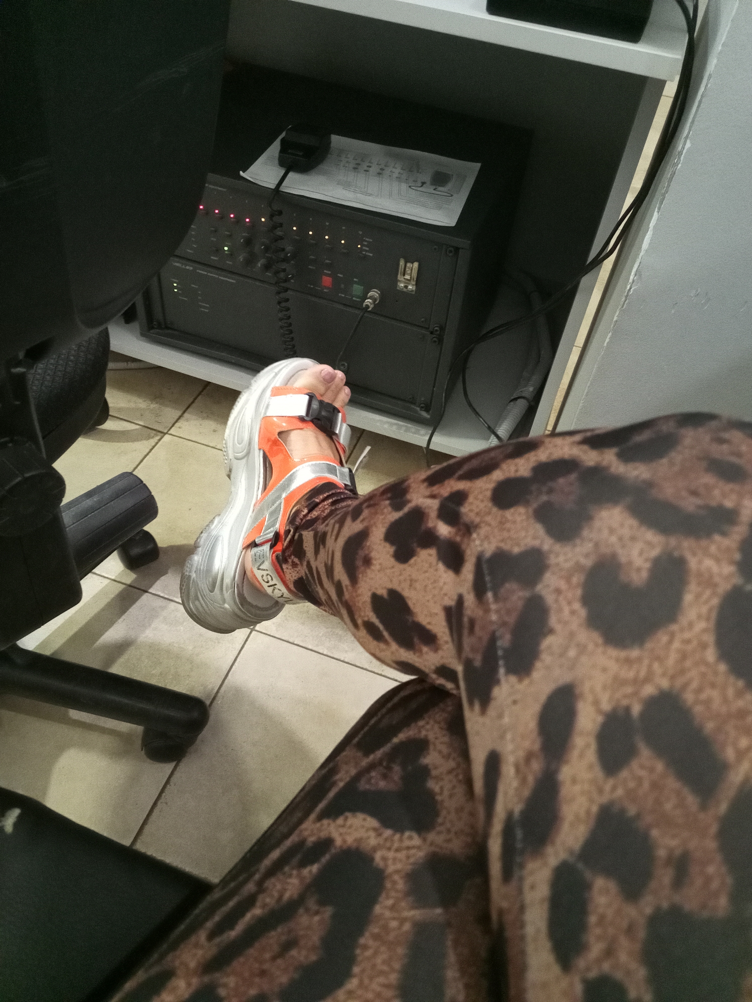 An overhead, medium shot shows a person's right leg and foot, which is wearing a leopard print legging and an orange and silver sandal, resting on a black office chair. The leg and foot are positioned in the foreground, taking up the right and lower portions of the frame. In the background, a black electronic device with several lights and knobs is visible, with a microphone attached to it. The device is placed on a white shelf, and a grey electrical cord runs along the shelf and down the wall. The floor appears to be tiled in a cream color, and the overall lighting suggests an indoor environment. The location is described as Unknown, Unknown.